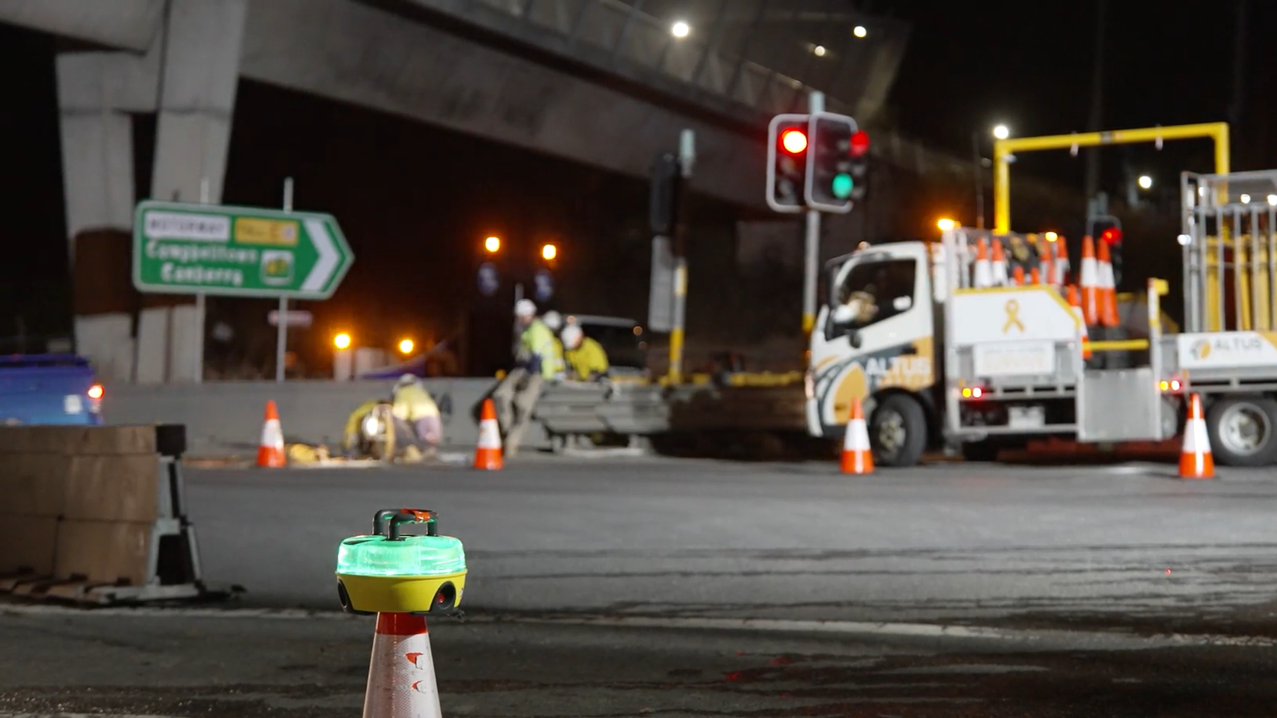 Nighttime construction scene on a highway with workers in safety gear, traffic cones, and a service vehicle near a red traffic light. Signage indicates directions to neighboring locations.