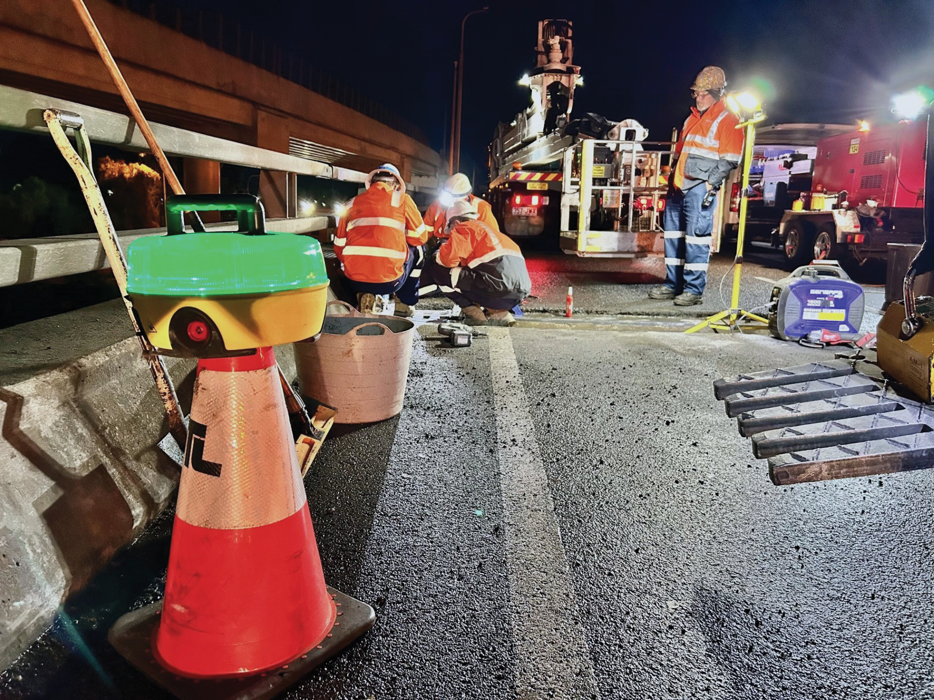 Workers in orange safety vests and helmets repairing a road at night, surrounded by traffic cones, equipment, and construction vehicles.