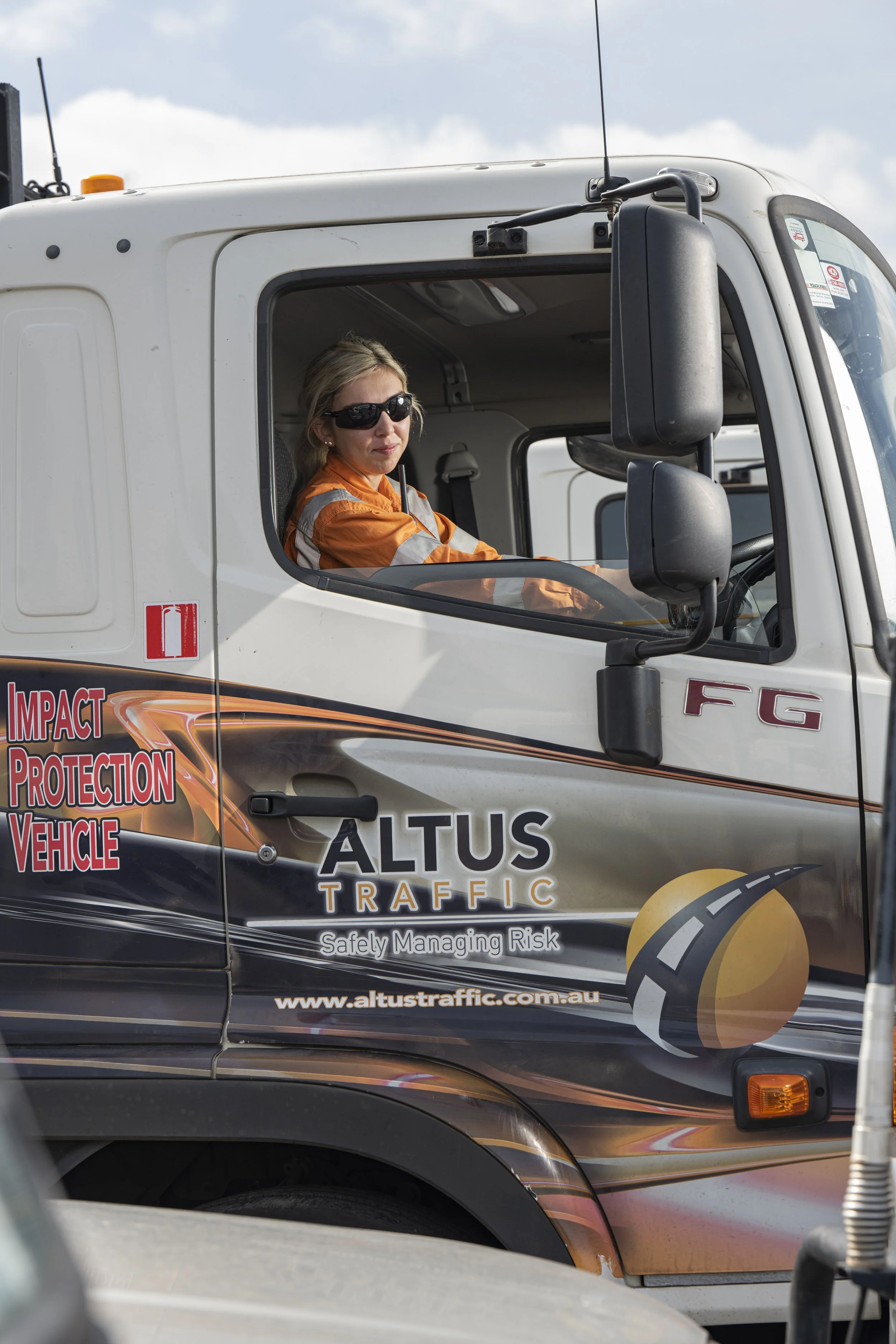 A woman wearing sunglasses and an orange safety jacket sitting in the driver's seat of a traffic management truck with 'Altus Traffic' branding.