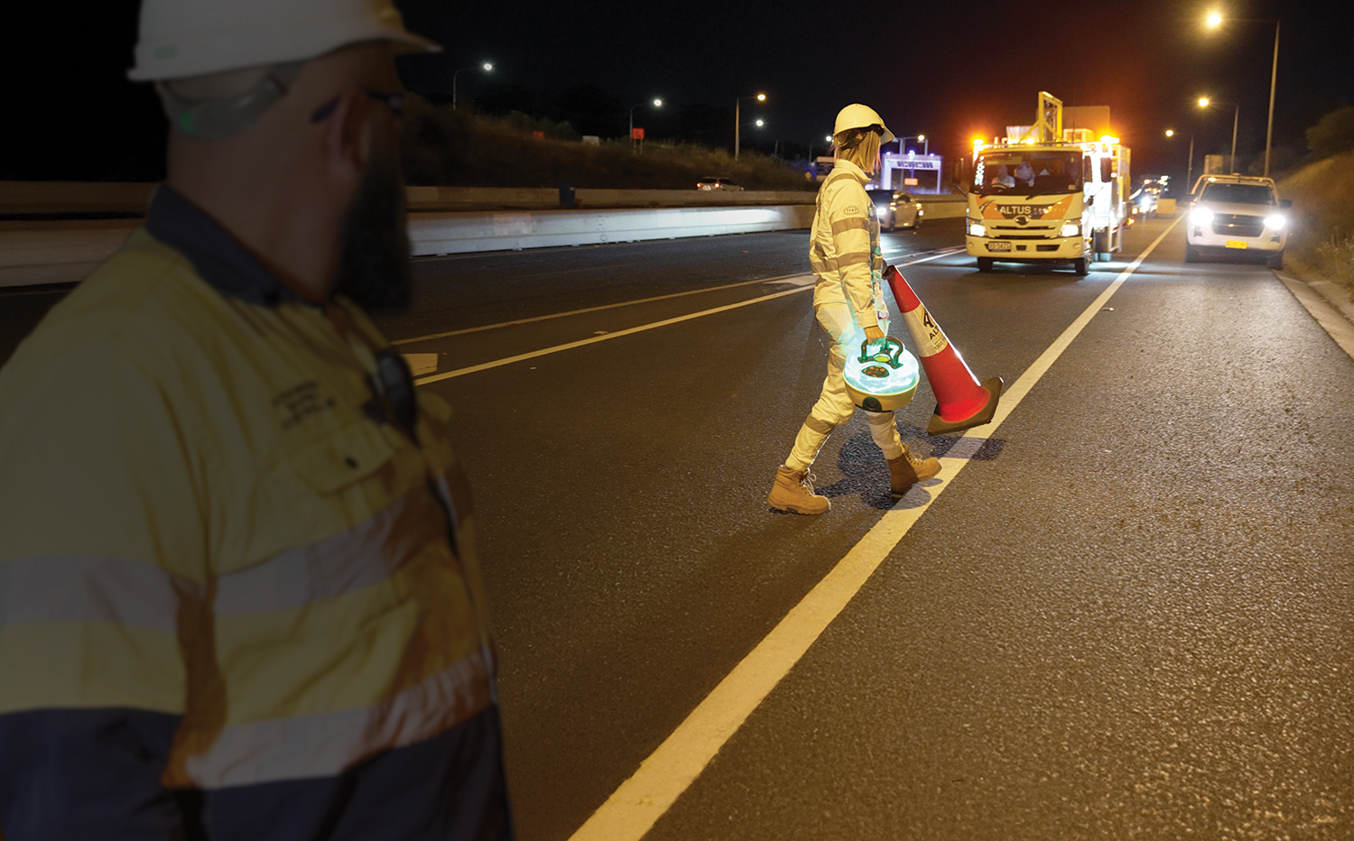 Workers in reflective vests and helmets performing road work at night with traffic in the background.
