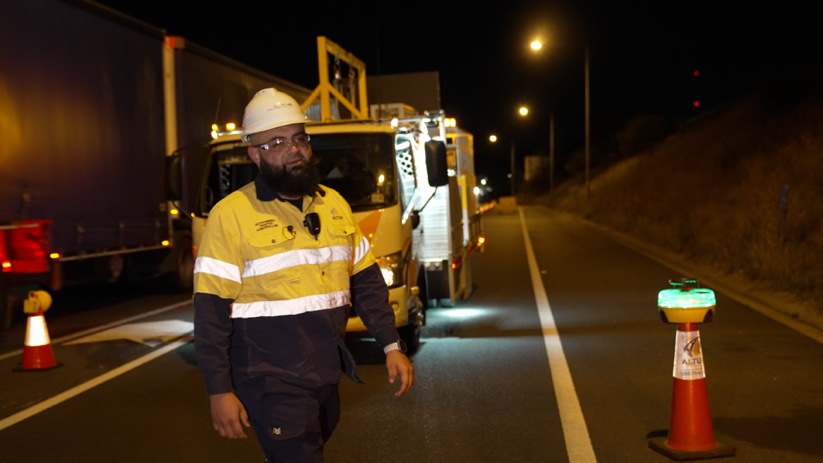 Nighttime scene on a highway with a worker in a yellow safety vest and white hard hat standing near a maintenance vehicle, surrounded by traffic cones, with trucks in the background and streetlights illuminating the area.
