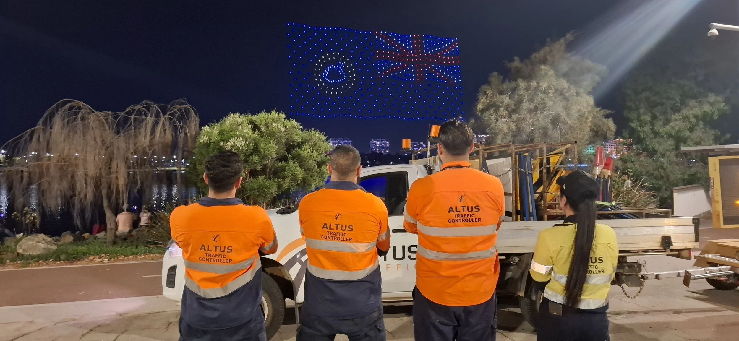 Four traffic controllers in orange and yellow vests standing in front of a Altus traffic control truck at night, with a large digital display showing a flag and a heart symbol.