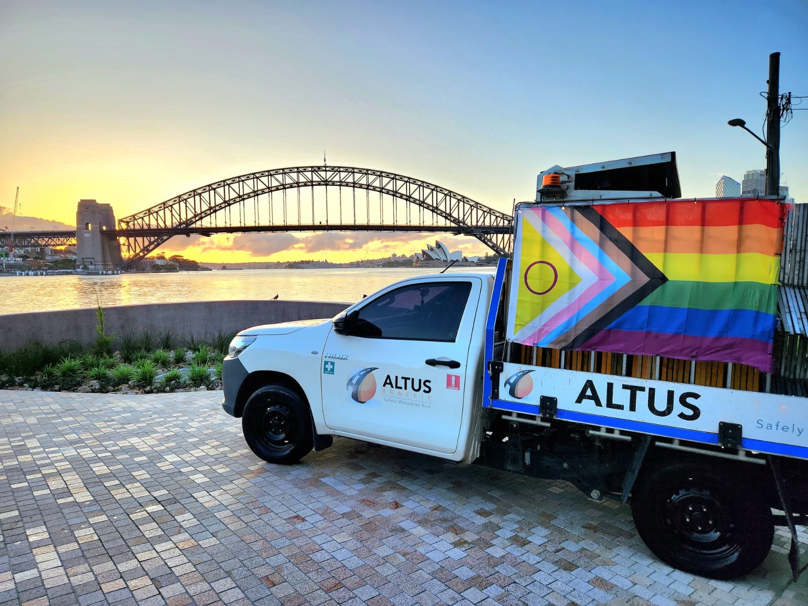 A white ALTUS traffic vehicle with rainbow pride flag parked near Sydney Harbour, with the Sydney Opera House and Harbour Bridge in the background at sunset.