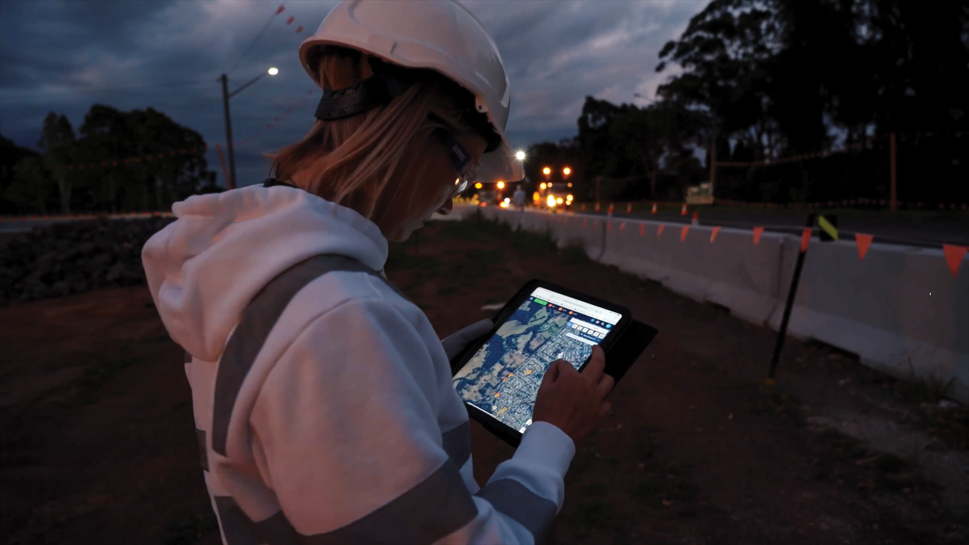 A woman wearing a white safety helmet and hoodie stands outdoors during dusk, using a tablet device with a digital map on the screen. She is near a construction site or roadwork area with traffic cones and barriers, with trees and a darkening sky in 