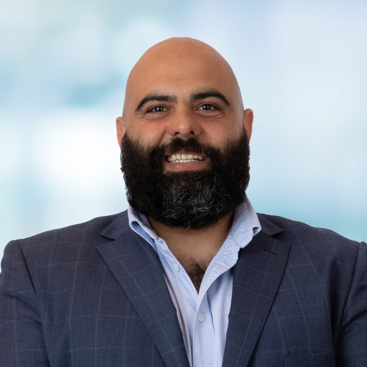 A man with a bald head and a thick, dark beard, smiling and wearing a dark plaid suit jacket and a light blue shirt against a blurred light blue background.