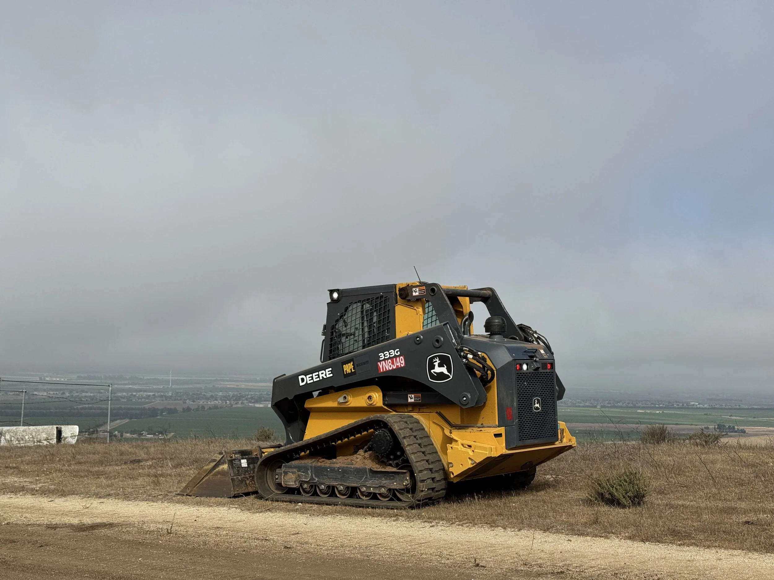 A yellow and black John Deere compact track loader construction vehicle on a dirt surface with open fields and a cloudy sky in the background.