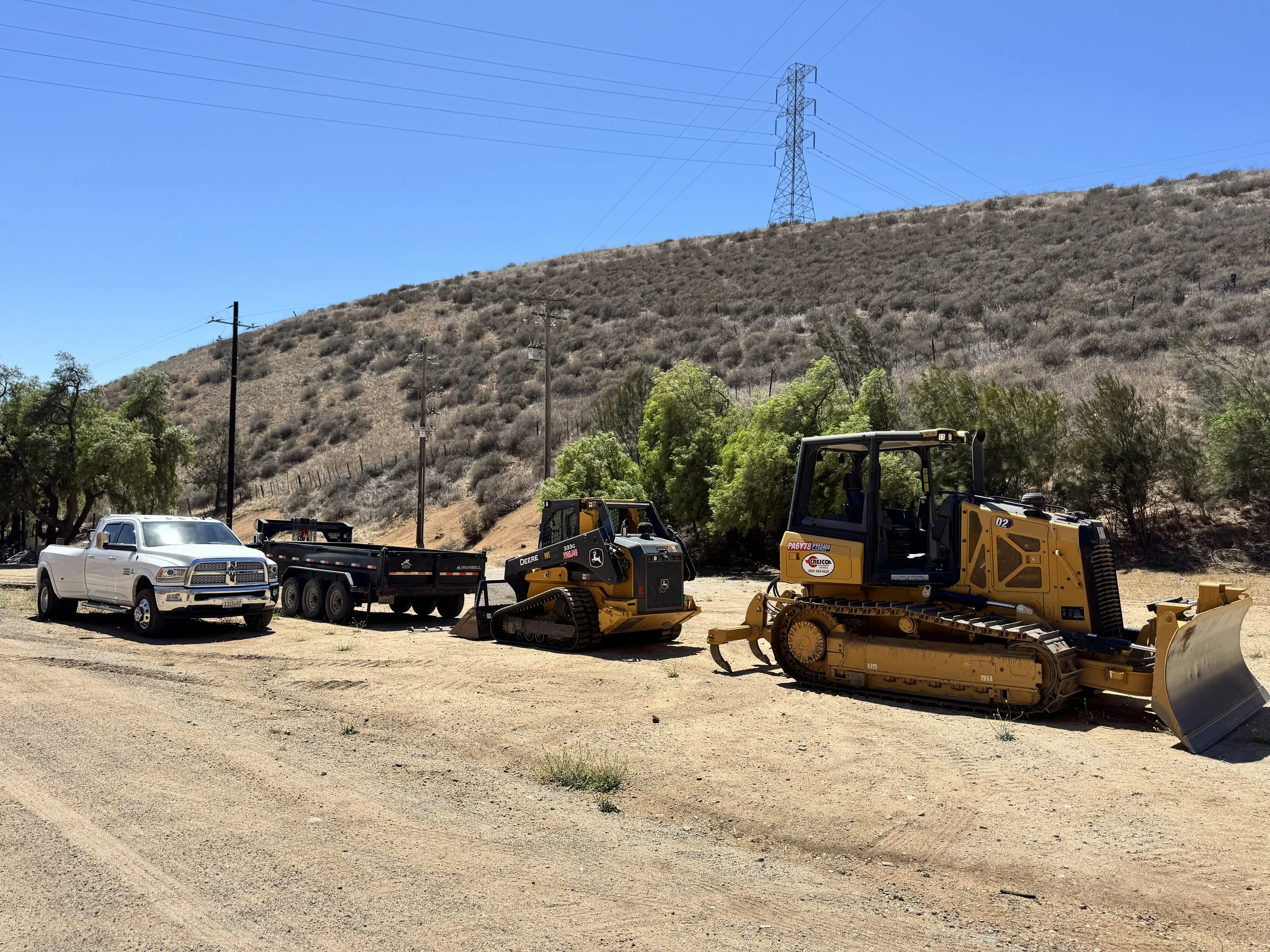 Construction vehicles including a white pickup truck, a black trailer, a small yellow bulldozer, and a larger yellow bulldozer parked on dirt in an outdoor area with a hillside and electrical towers in the background.