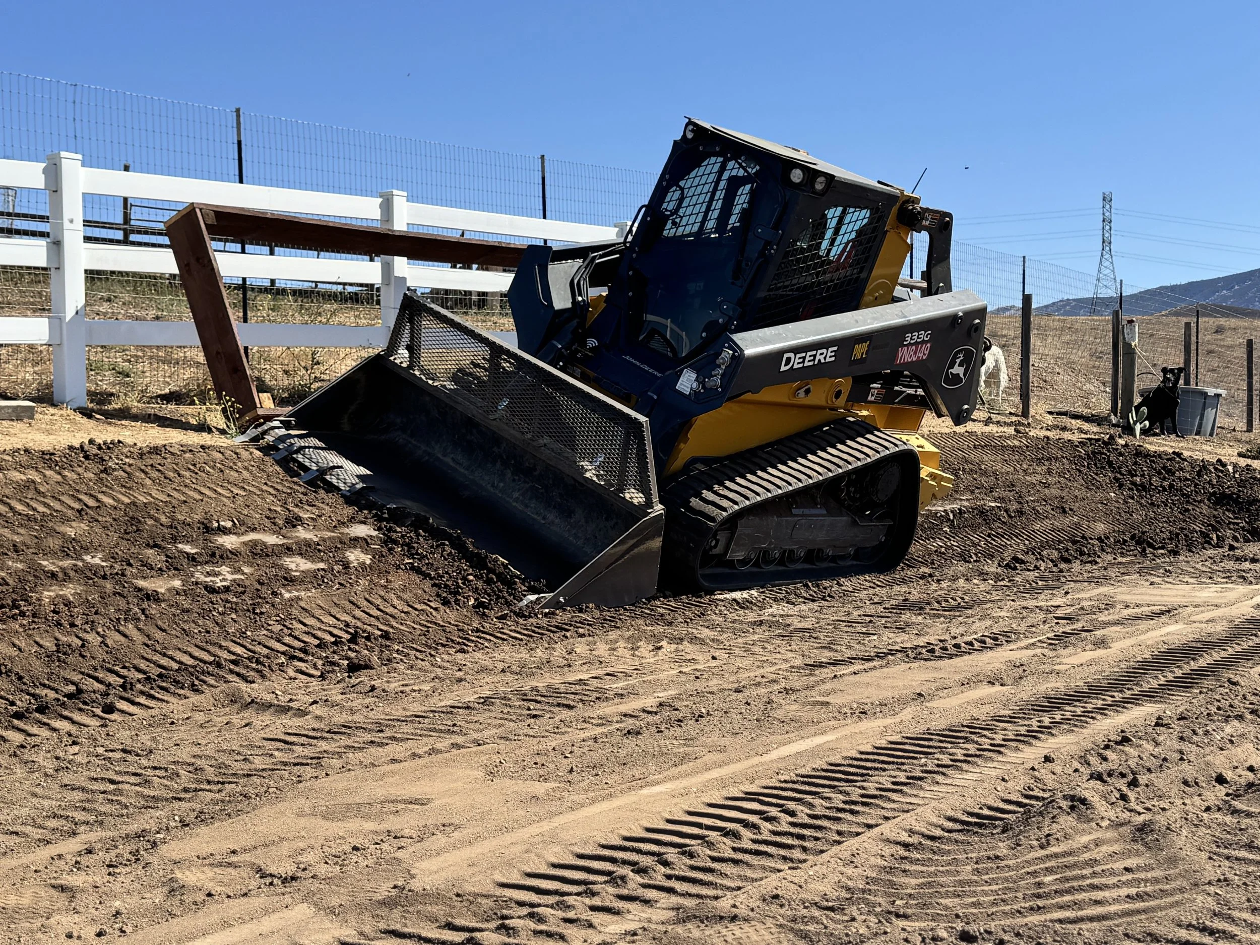 A yellow and black John Deere compact track loader pushing dirt on a construction site with a white fence and hills in the background.