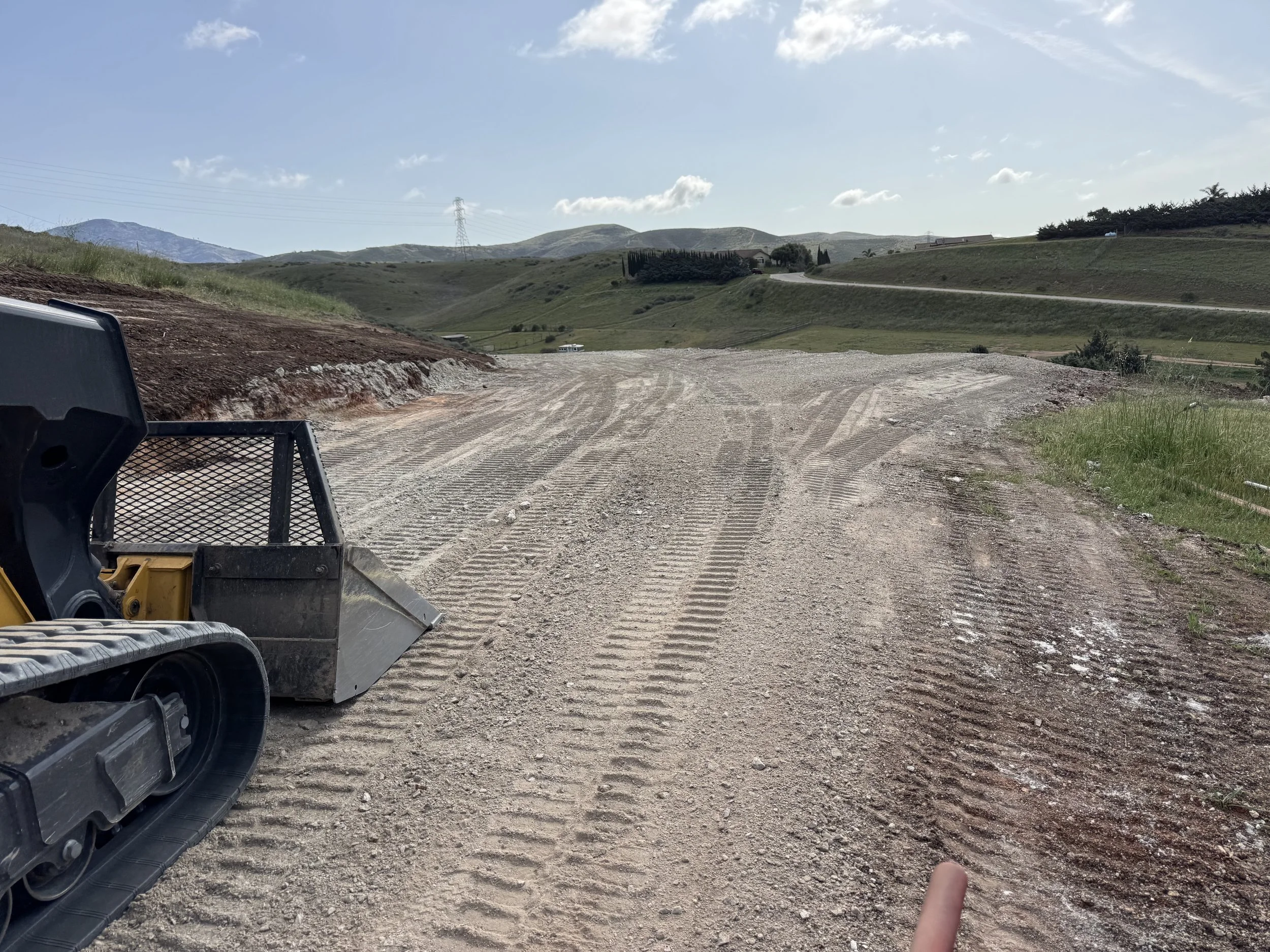 Dirt road under construction with construction equipment on the left, grassy hills, and a partly cloudy sky in the background.