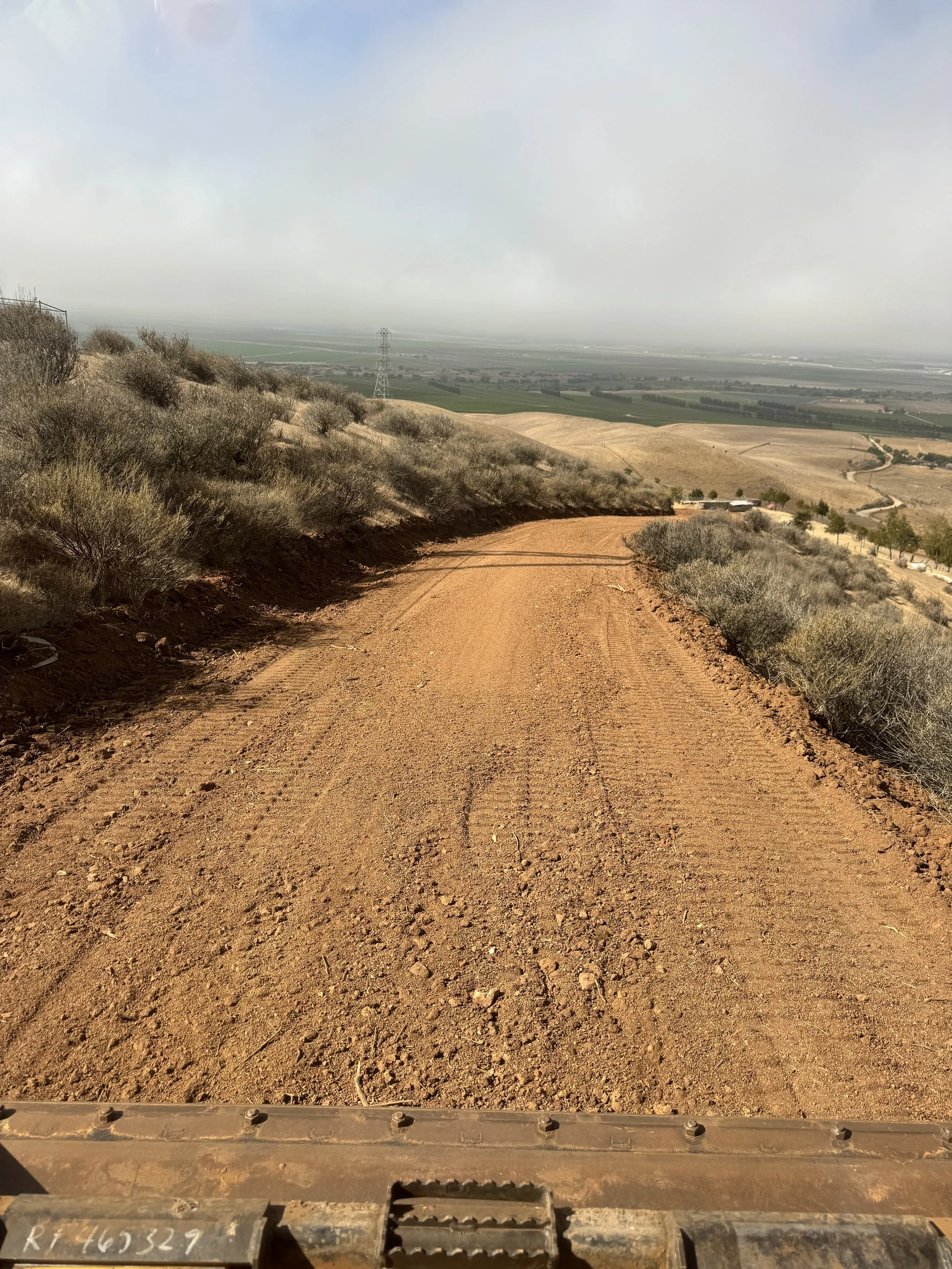 View from the front of an off-road vehicle on a dirt trail in a hilly desert landscape with scrub vegetation, distant agricultural fields, and a cloudy sky.