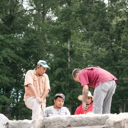 Four young men on a rocky ledge outdoors surrounded by green trees, one sitting, one standing, and two leaning over, engaged in an activity together.