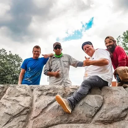 Four men posing outdoors on a stone wall, with cloudy sky and trees in the background.