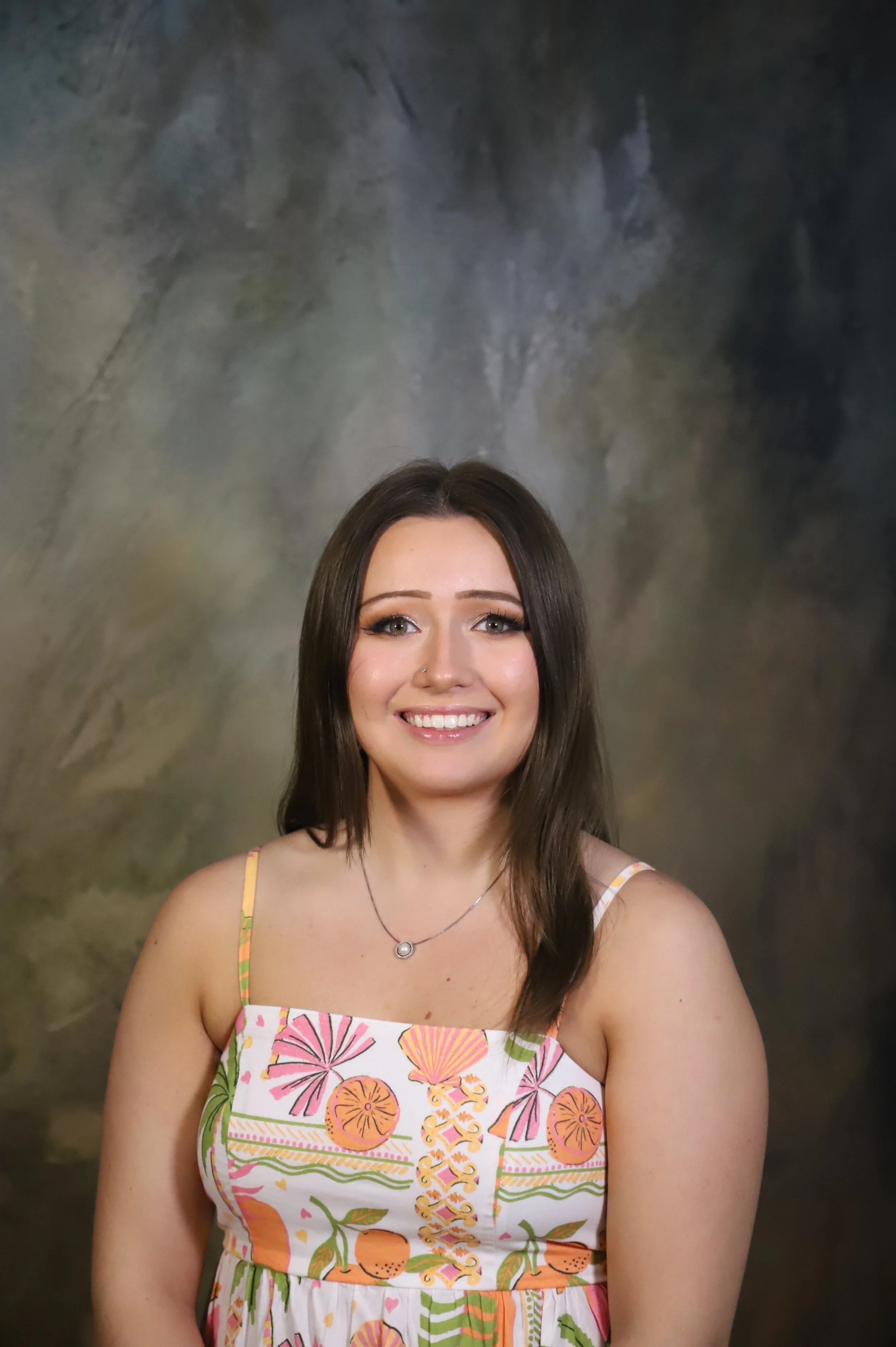 A woman with long dark hair smiling, wearing a colorful summer dress with floral and fruit patterns, and a small necklace, against a dark abstract background.