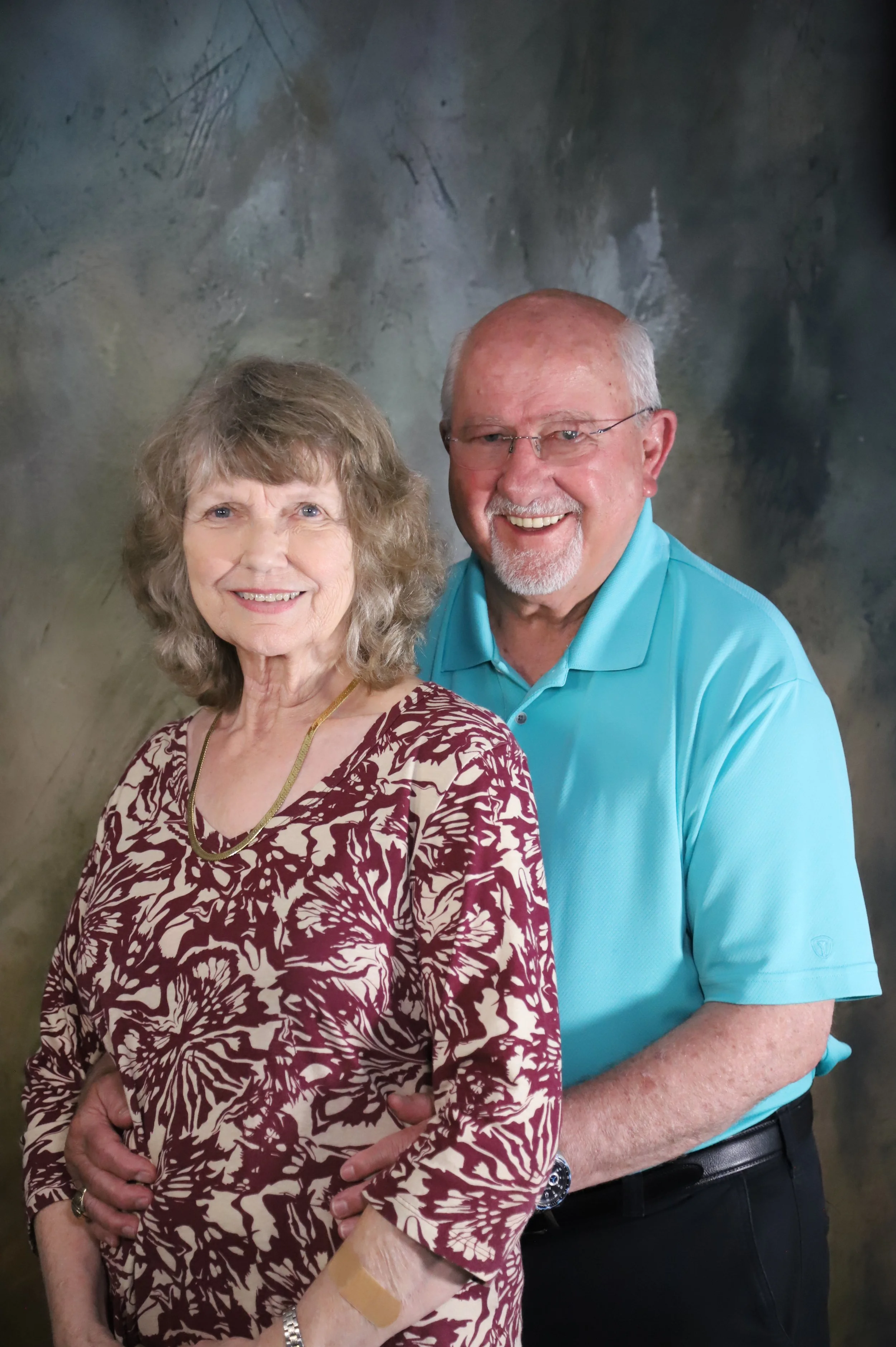 An elderly woman with curly light brown hair wearing a maroon and beige floral dress and a gold necklace, standing in front of an elderly man with glasses, short white hair, beard, wearing a turquoise polo shirt and black pants, both smiling.