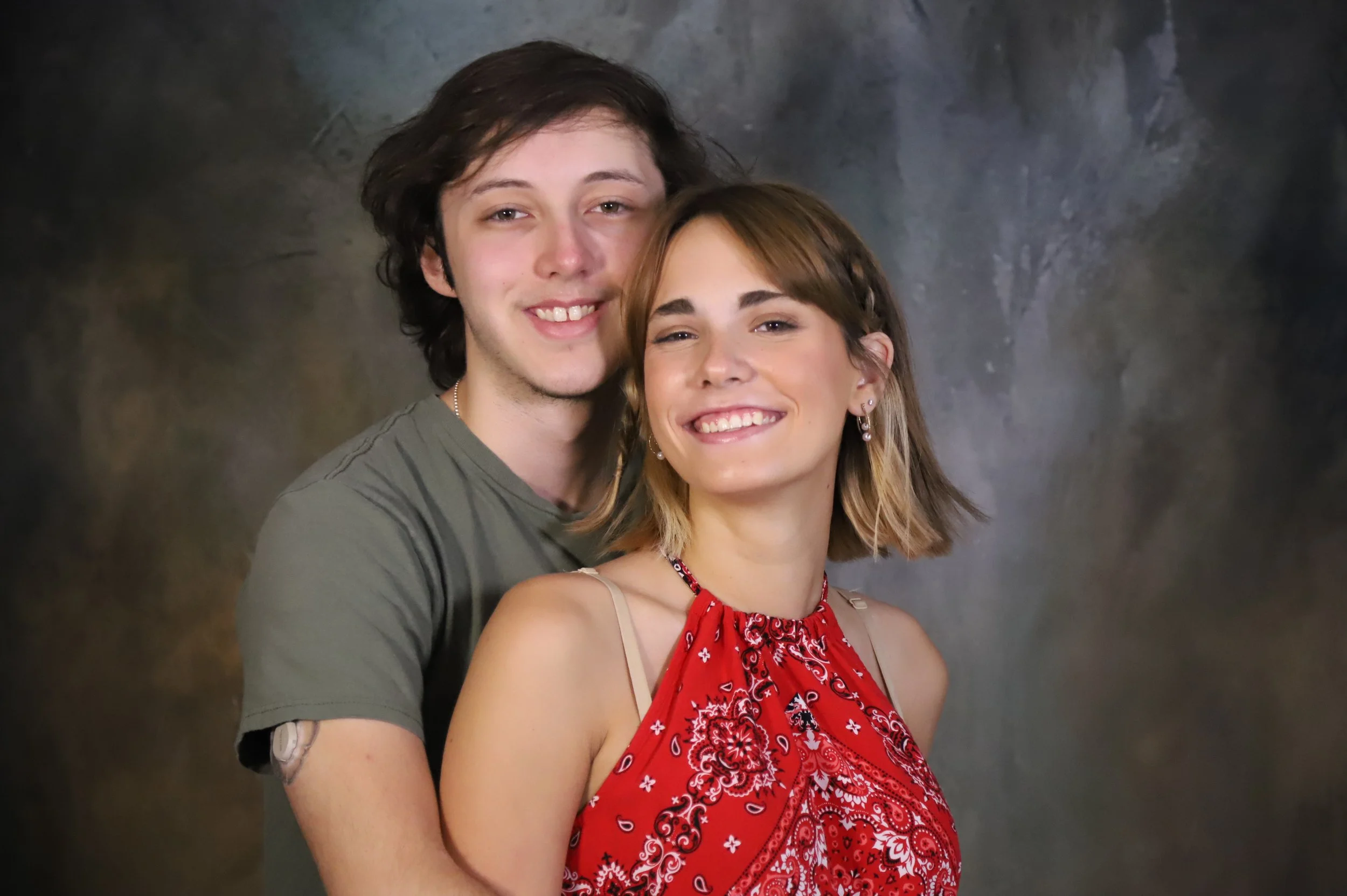 A young man with dark hair and a young woman with light brown hair and earrings, smiling and standing close together against a dark, blurred background.