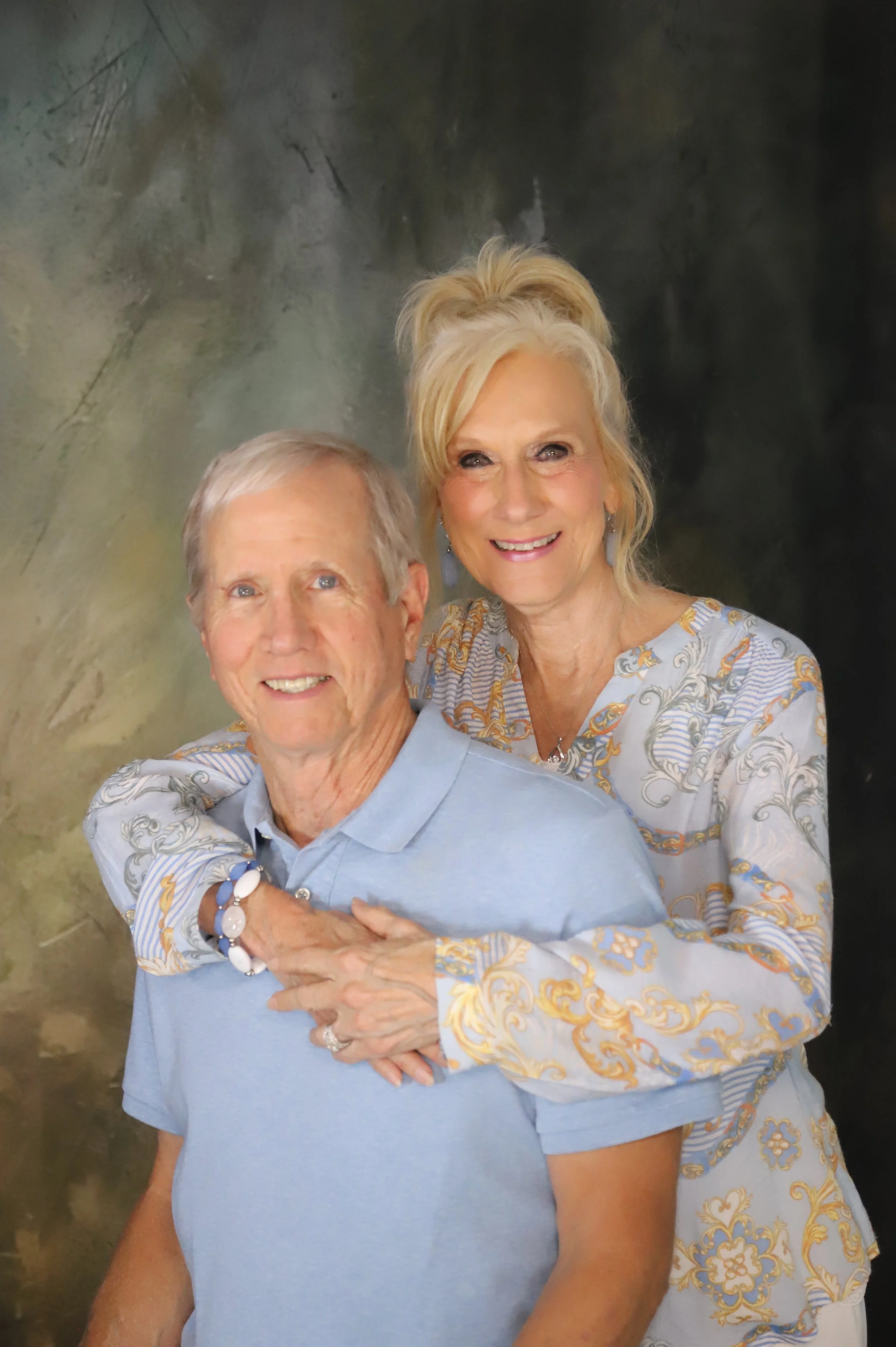 A smiling older man and woman pose together, with the woman embracing the man from behind on a dark, painterly background.