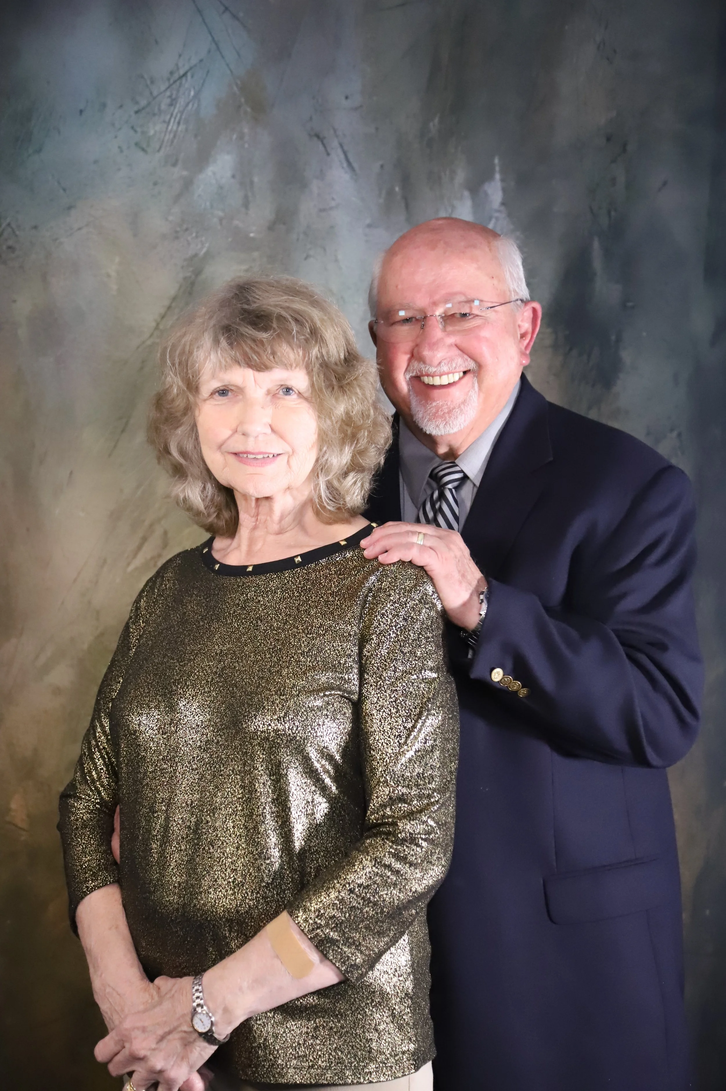 An elderly woman in a shiny gold and black dress standing beside an elderly man in a dark suit and tie, on a painted studio backdrop.