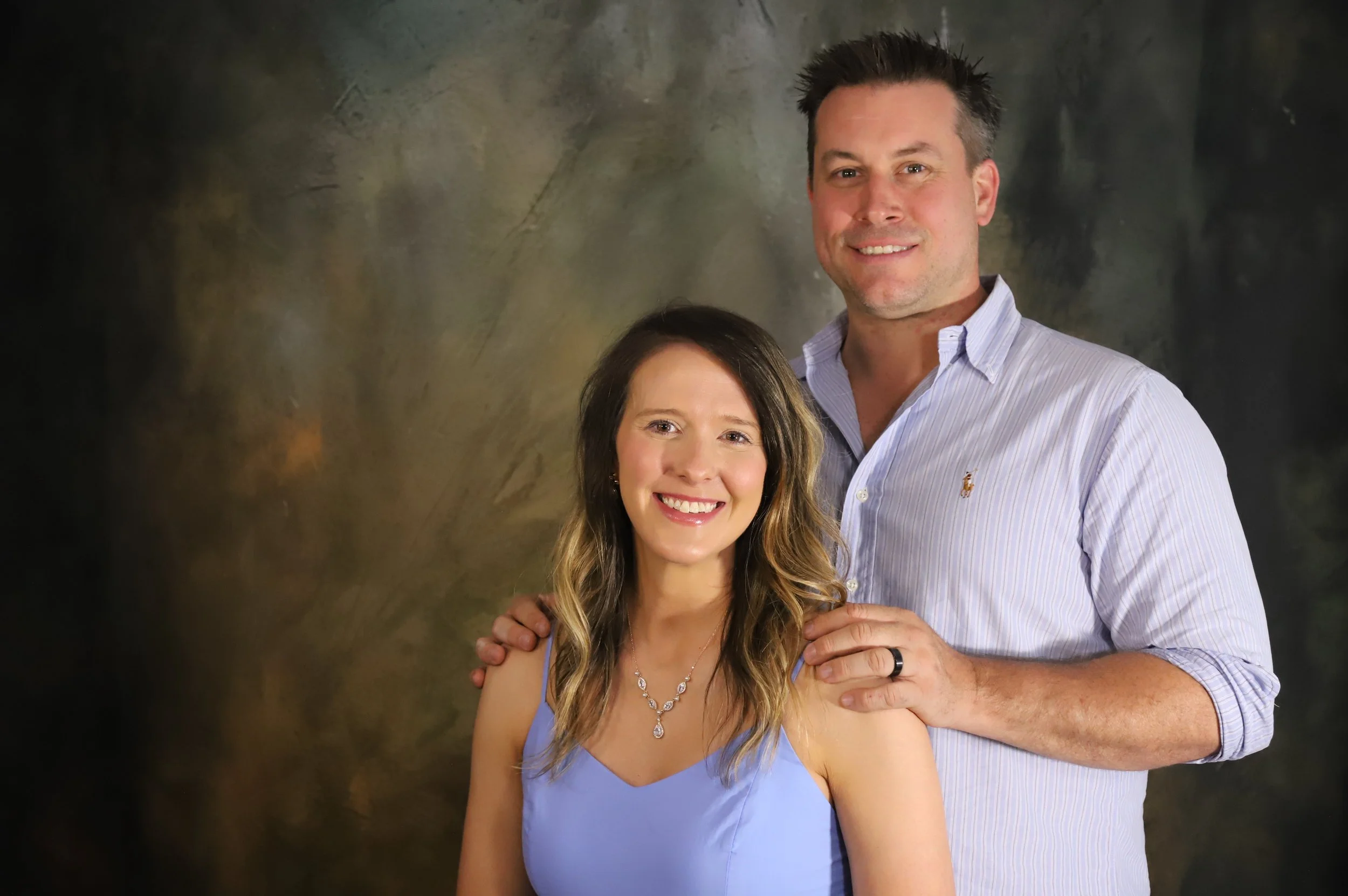 A smiling couple posing for a portrait. The woman has long, wavy hair and is wearing a light blue sleeveless dress with a necklace. The man has short dark hair, is wearing a light blue striped button-down shirt, and has his hand on the woman's should