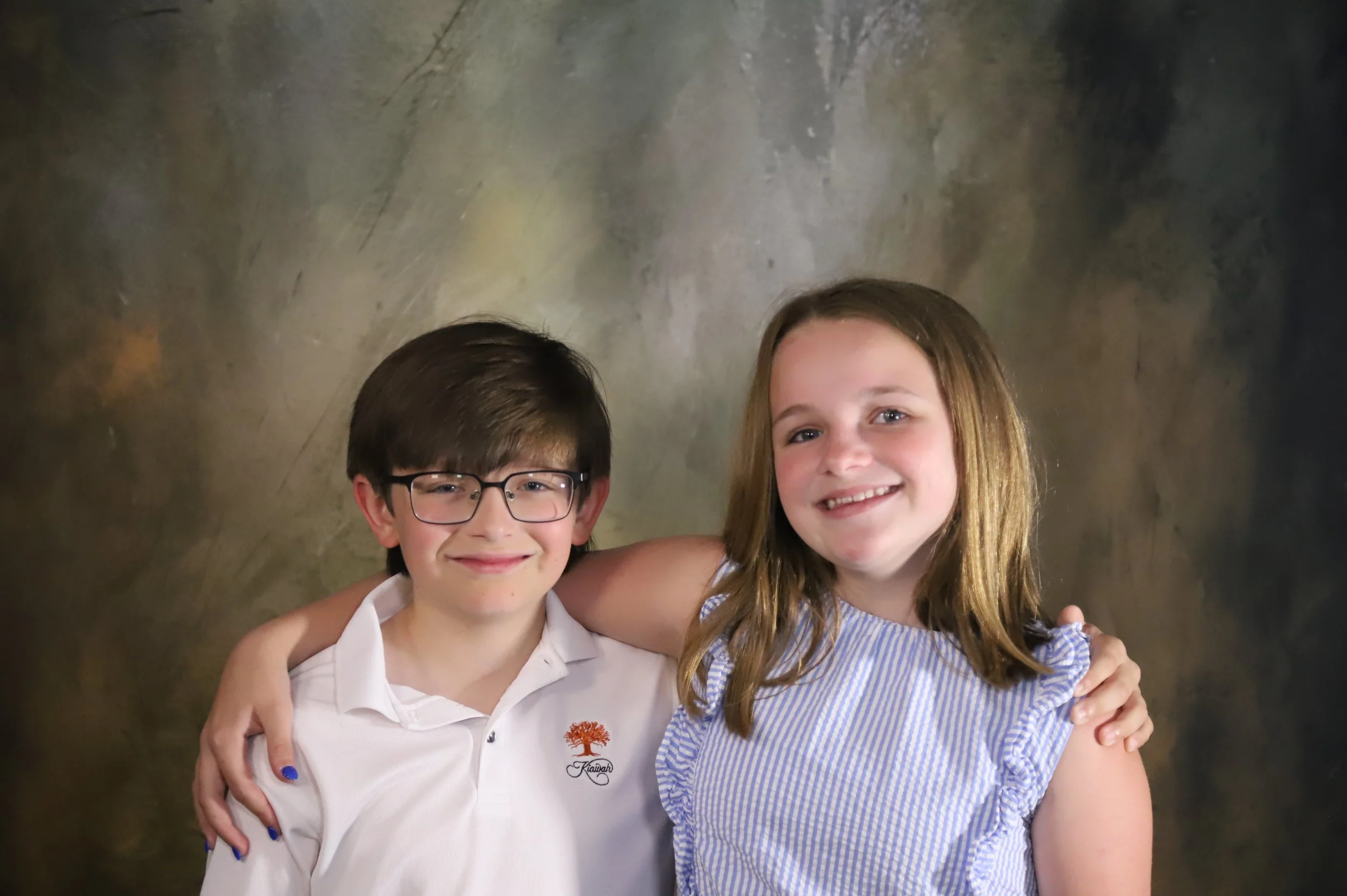 A boy and girl with their arms around each other smiling at the camera, standing against a blurred background.
