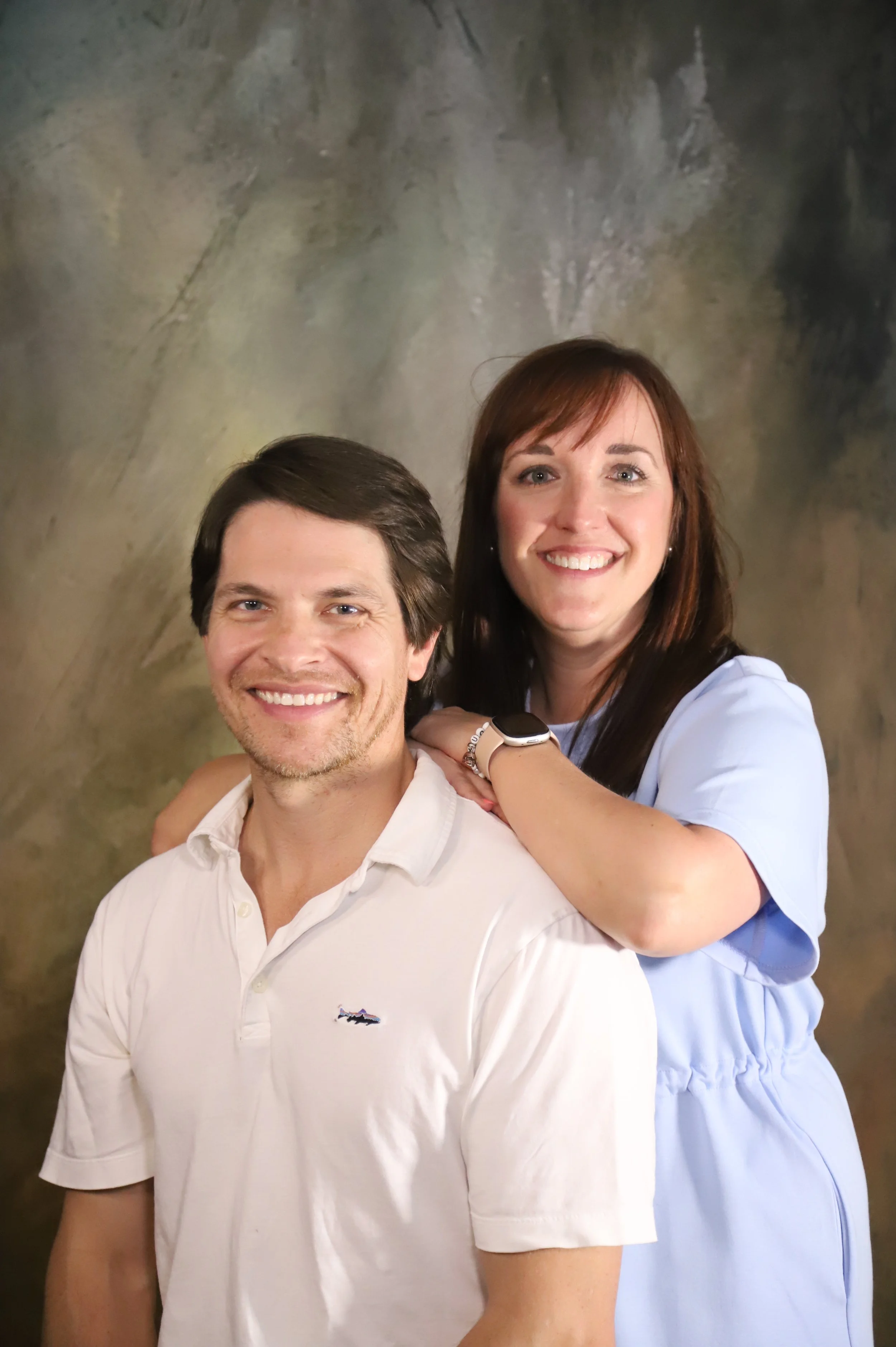 A smiling couple posing together, with the woman standing behind and resting her arms on the man's shoulders. The woman has shoulder-length brown hair and is wearing a light blue dress with a smartwatch on her wrist. The man has dark brown hair and i