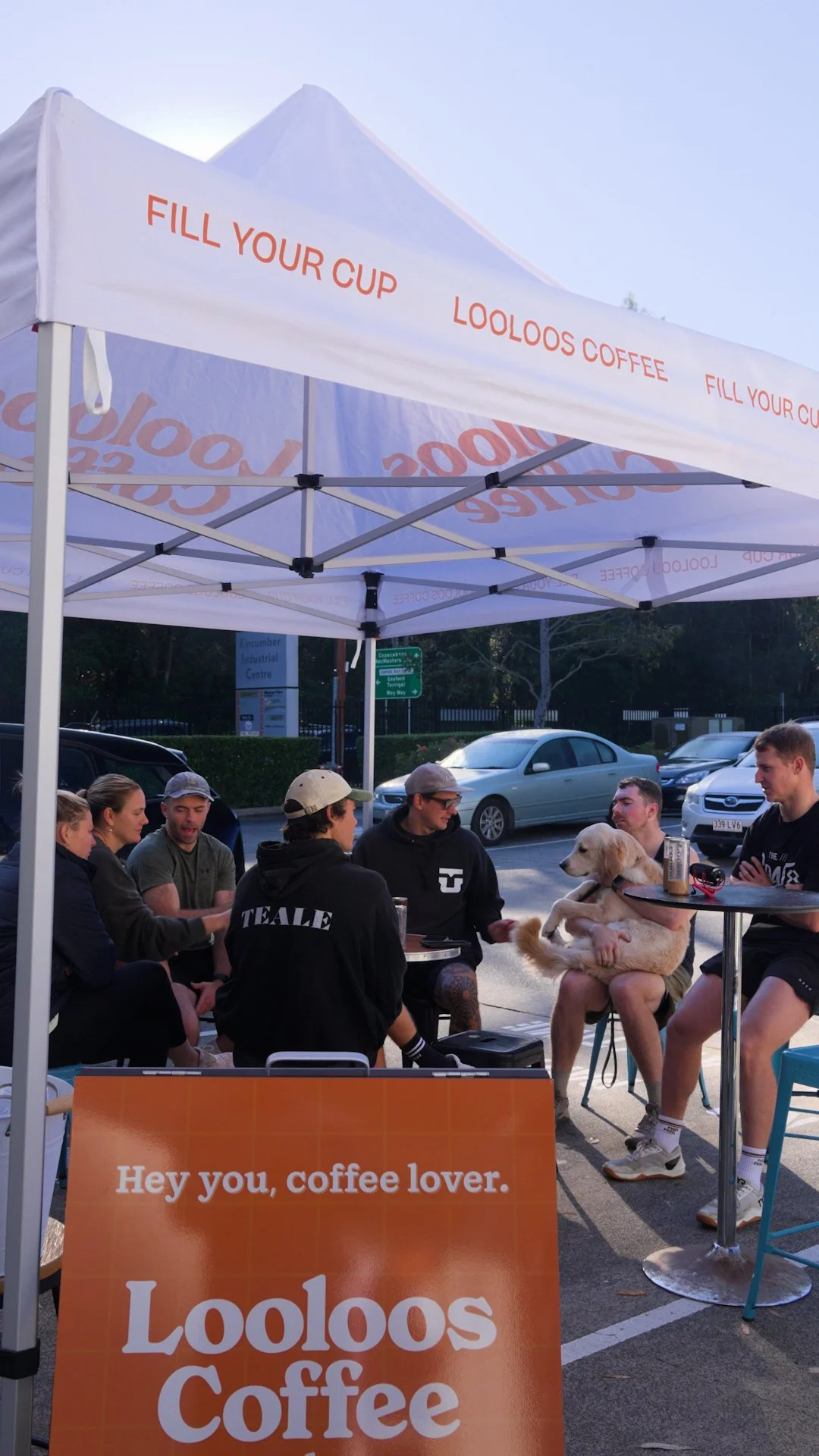 People sitting at a coffee stand under a white canopy with orange lettering, with cars parked behind them. A sign in front advertising Lolooos Coffee.