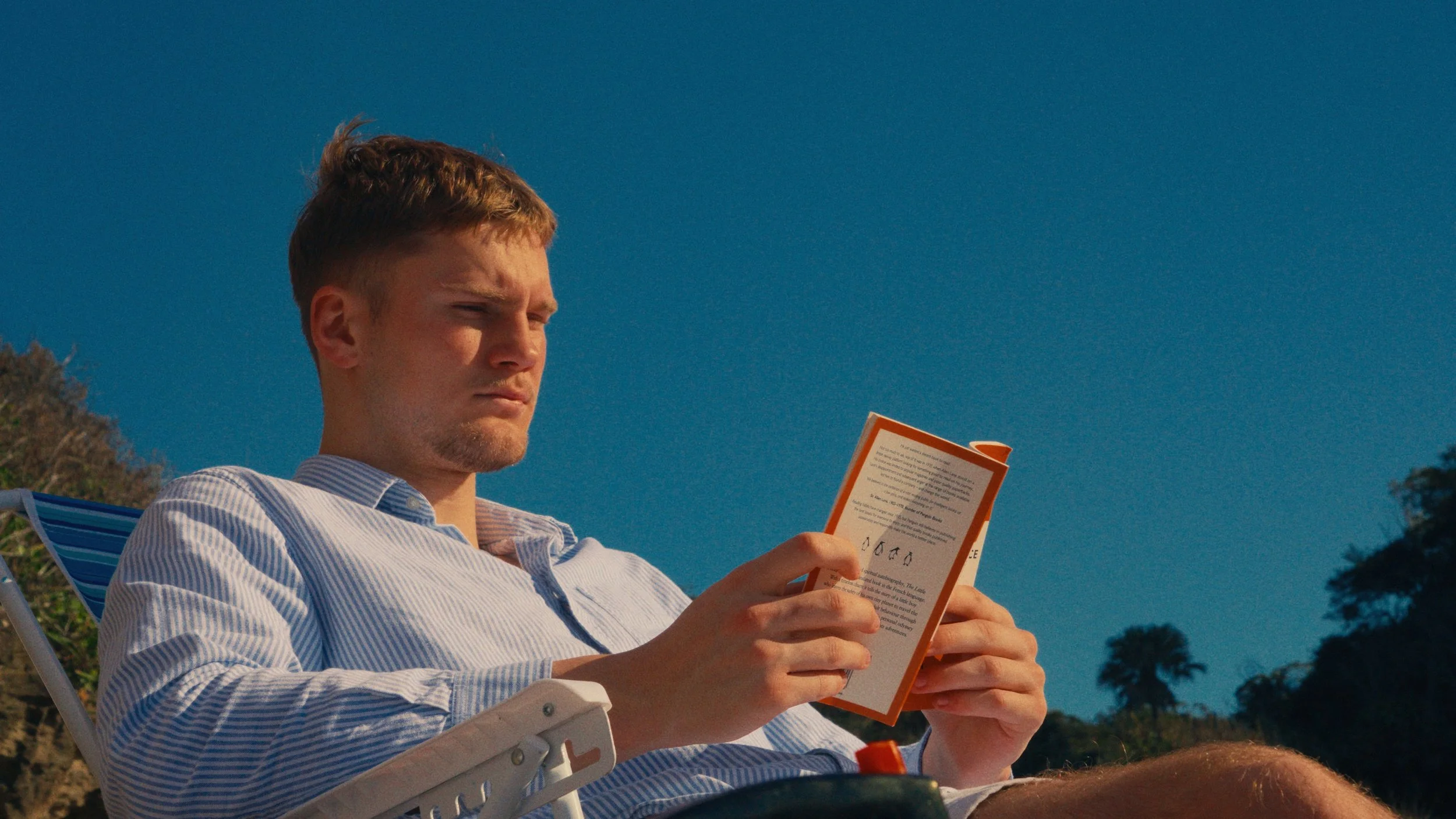 A young man with light brown hair and a striped shirt sitting outdoors and reading a booklet under a clear blue sky.