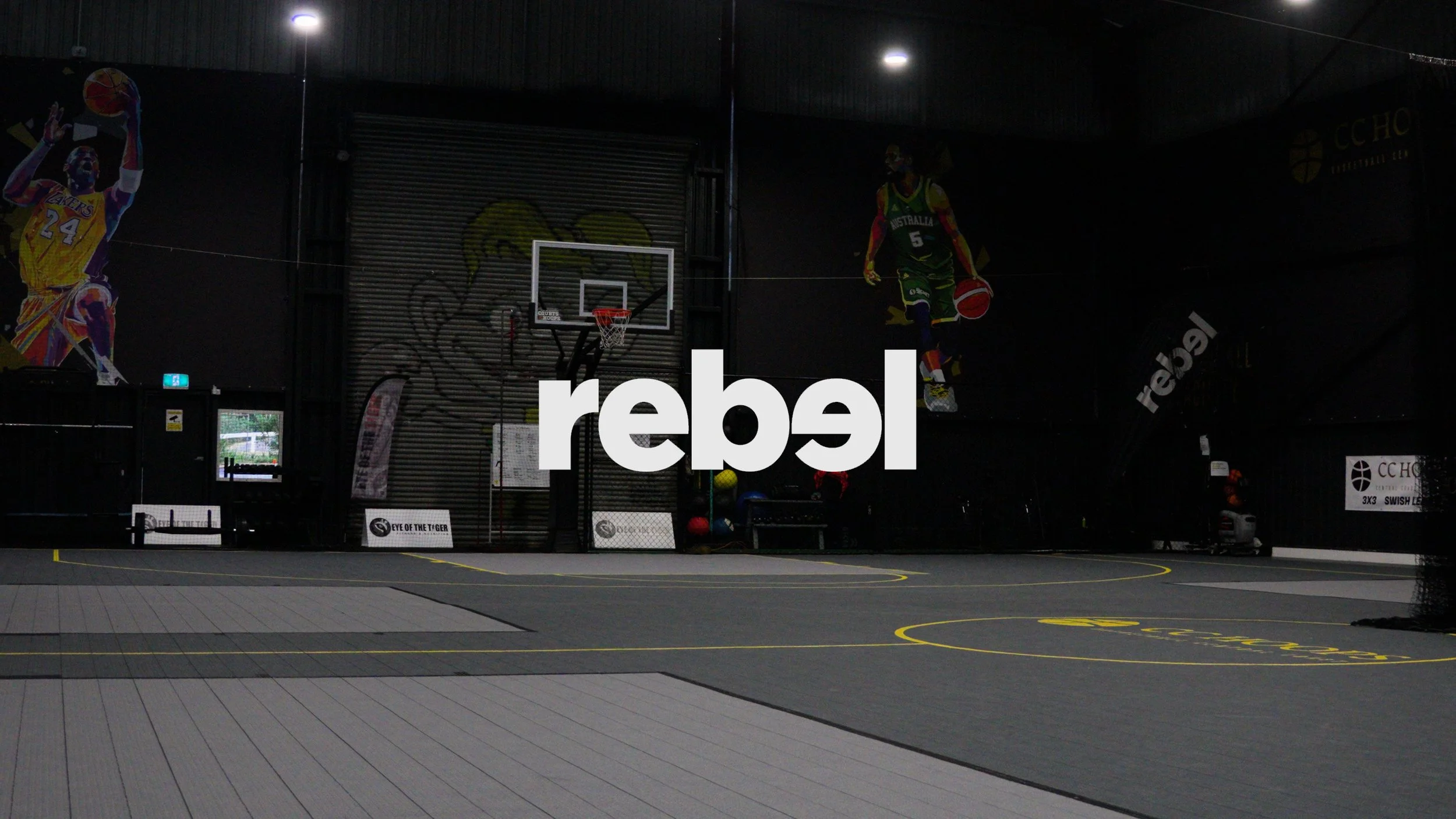Indoor sports court with black walls, murals of basketball players, and a large Rebel logo overlay.