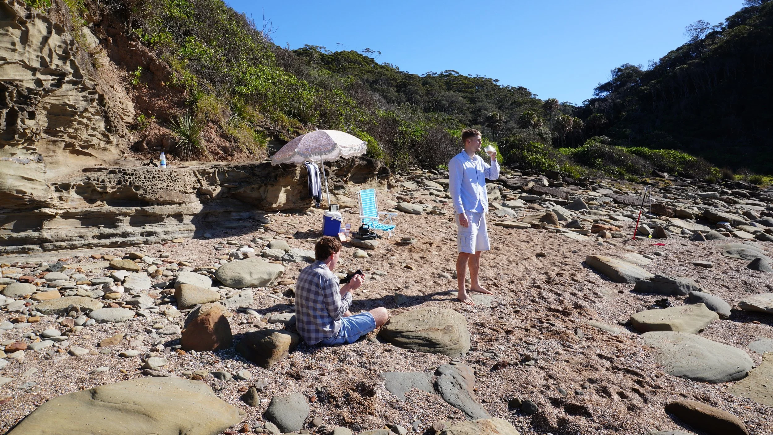 Two young men on a rocky beach with a clothesline, beach umbrella, and lounge chair, one standing drinking from a glass and the other sitting on rocks, with dense green vegetation and blue sky in the background.