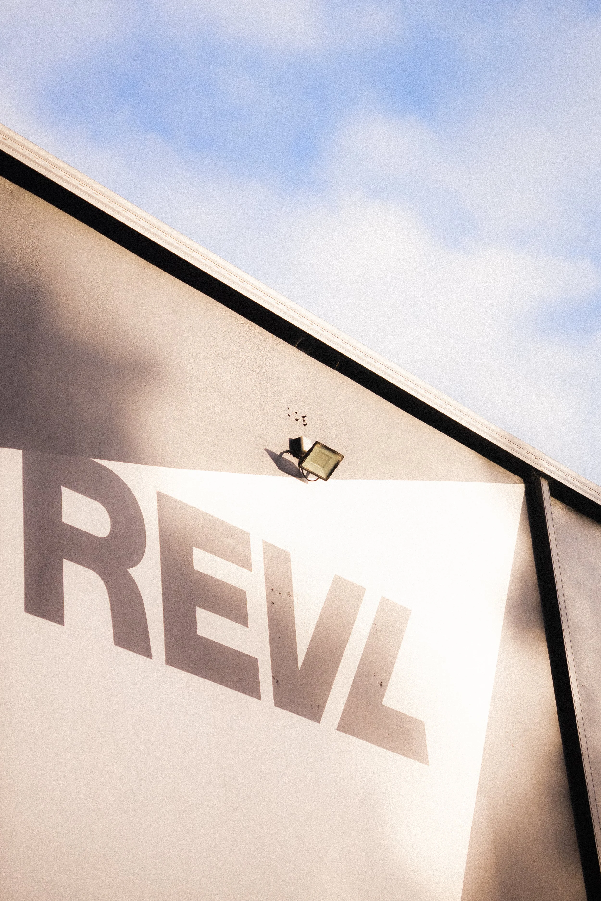 Close-up of a building with a large faded sign spelling 'REVL', a floodlight above, and a blue sky with light clouds in the background.