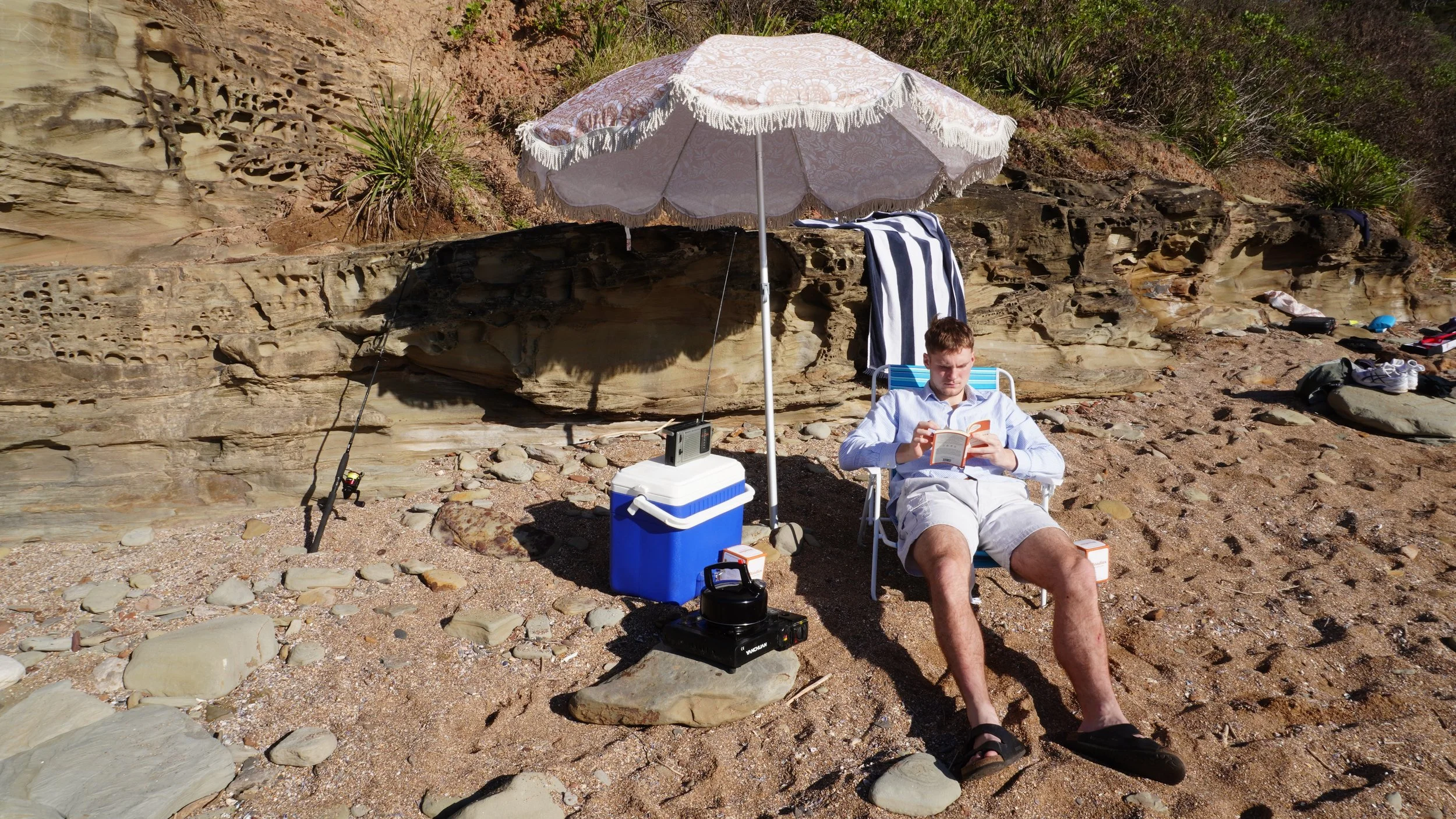 A man sitting on a beach chair reading a book under a large white umbrella with a cooler, radio, and fishing rod nearby, on a rocky sandy beach with rock formations and sparse vegetation in the background.