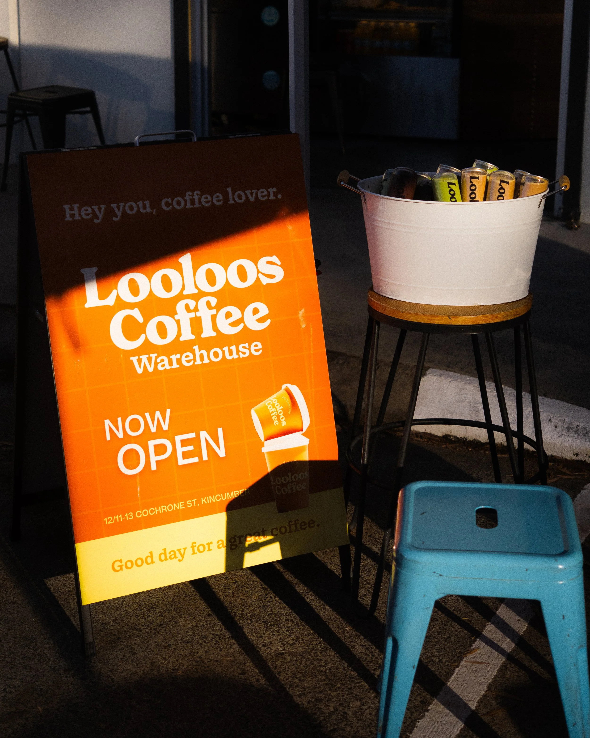 Signboard for Looloos Coffee Warehouse with a bucket of cans and a blue stool outside a cafe at dusk.