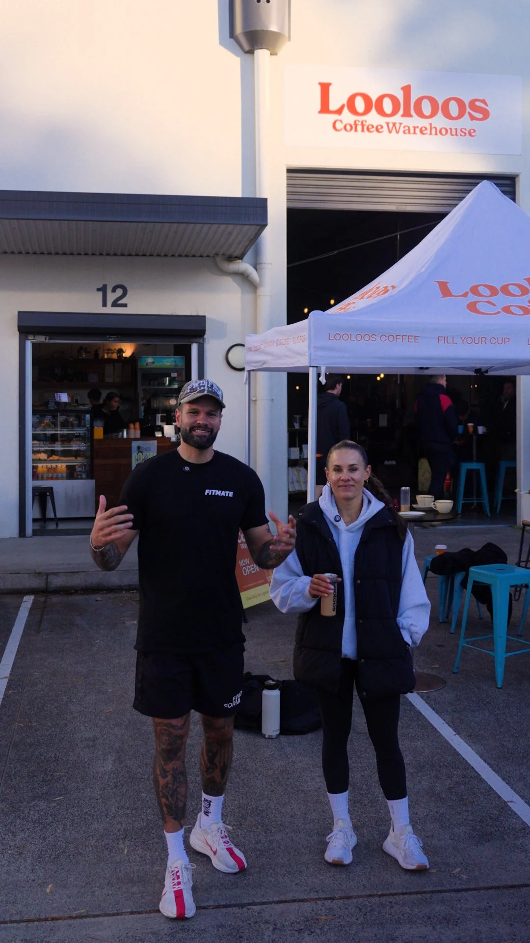 Two people standing outside a coffee shop called Lootos Coffee Warehouse, holding drinks, with the shop sign and a white canopy tent visible.