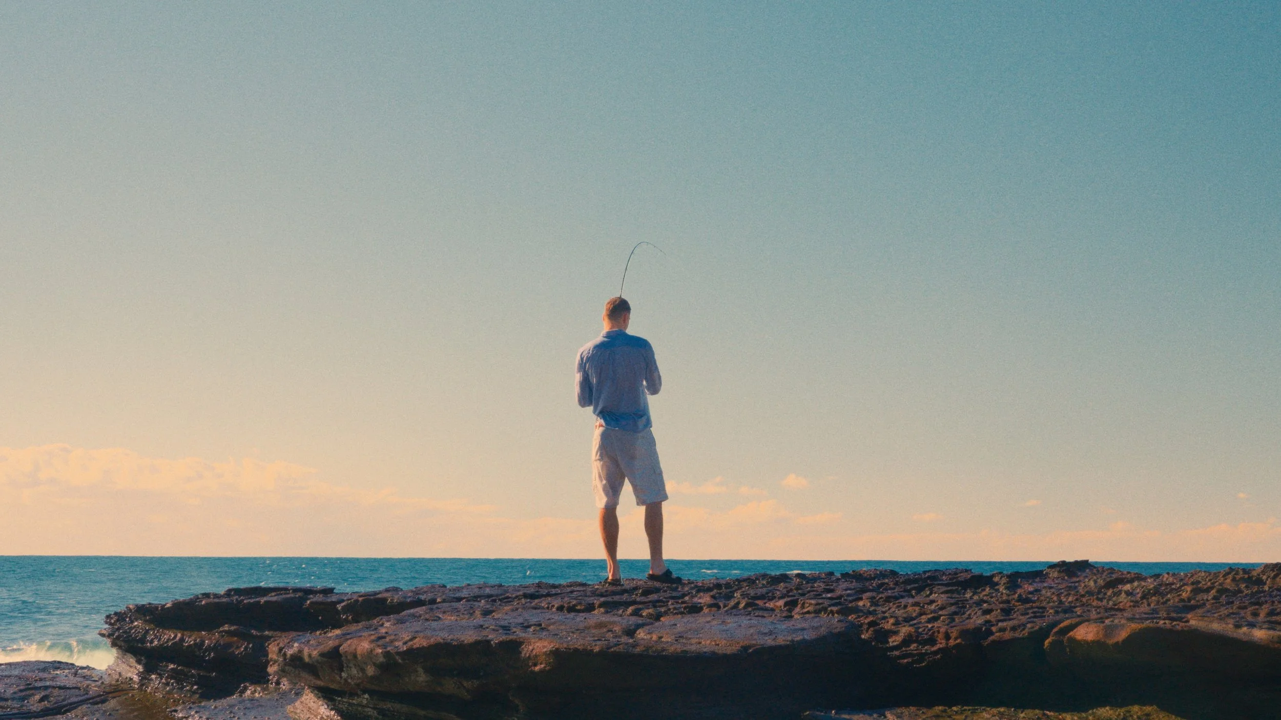 A man standing on rocky shoreline fishing with a fishing rod in the ocean during sunset or sunrise.