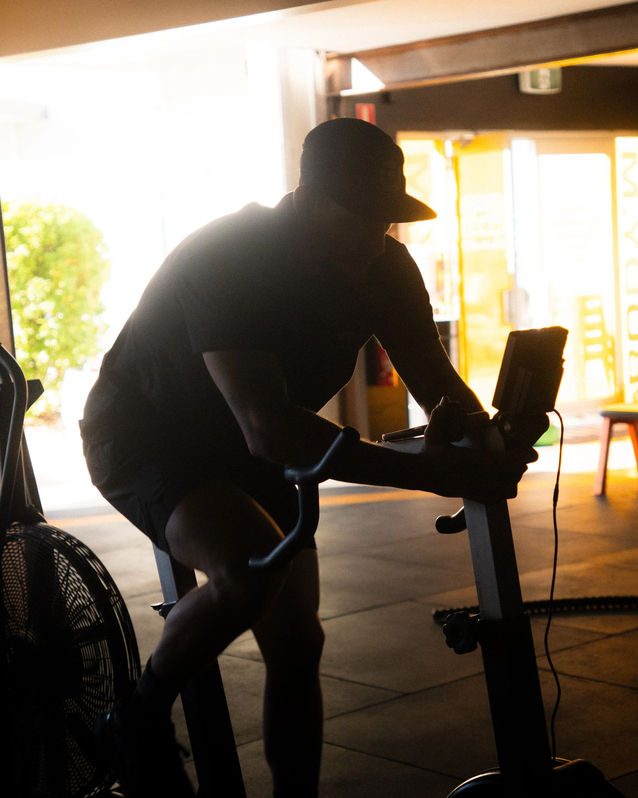 A person silhouetted on a stationary exercise bike inside a gym, with sunlight coming through the windows behind them.