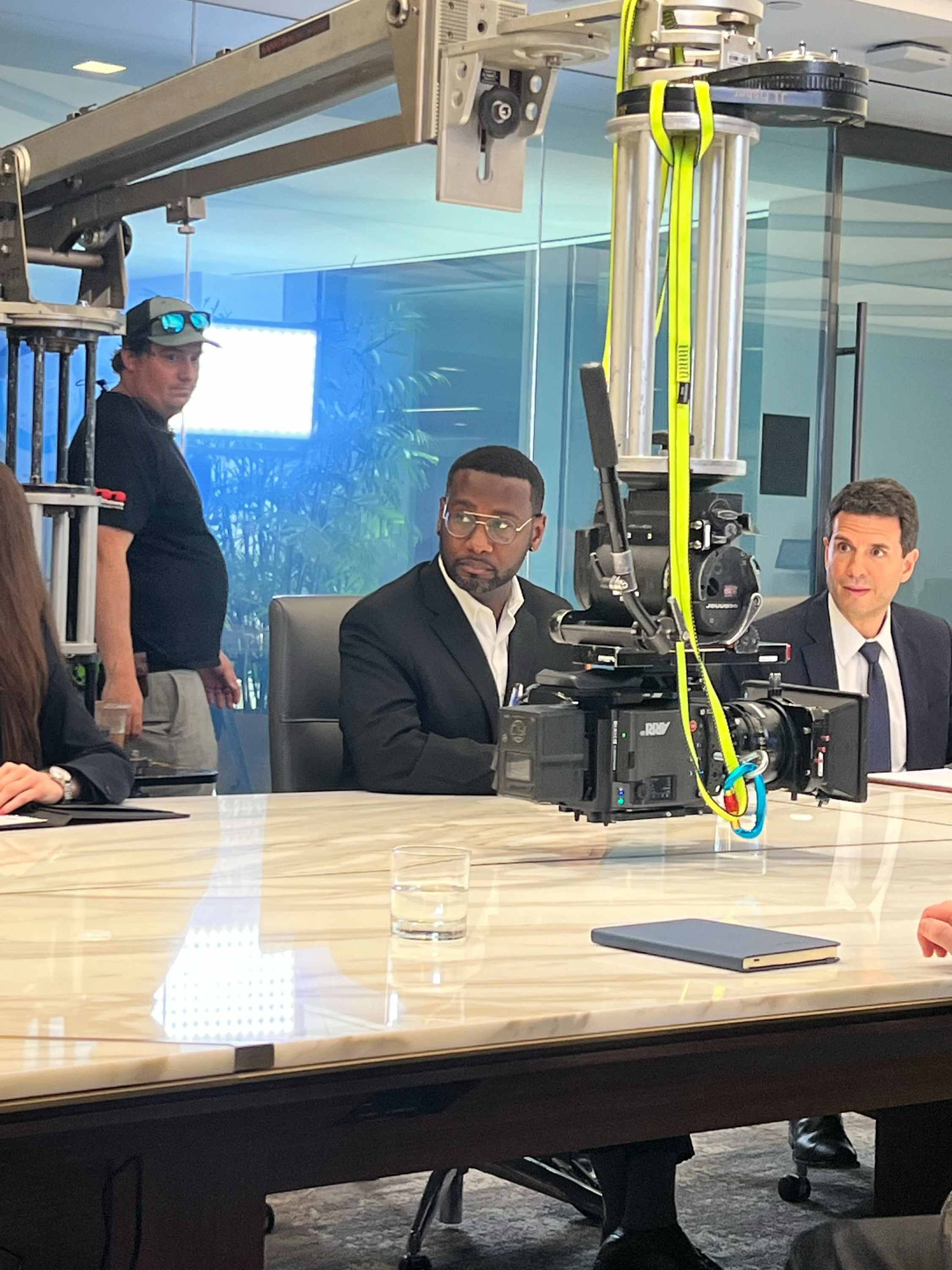 A camera crane is set up above a conference table with three men, one of whom is seated and wearing glasses and a black suit. There is a glass of water and a closed notebook on the table. A person with sunglasses and a hat is standing behind them, an