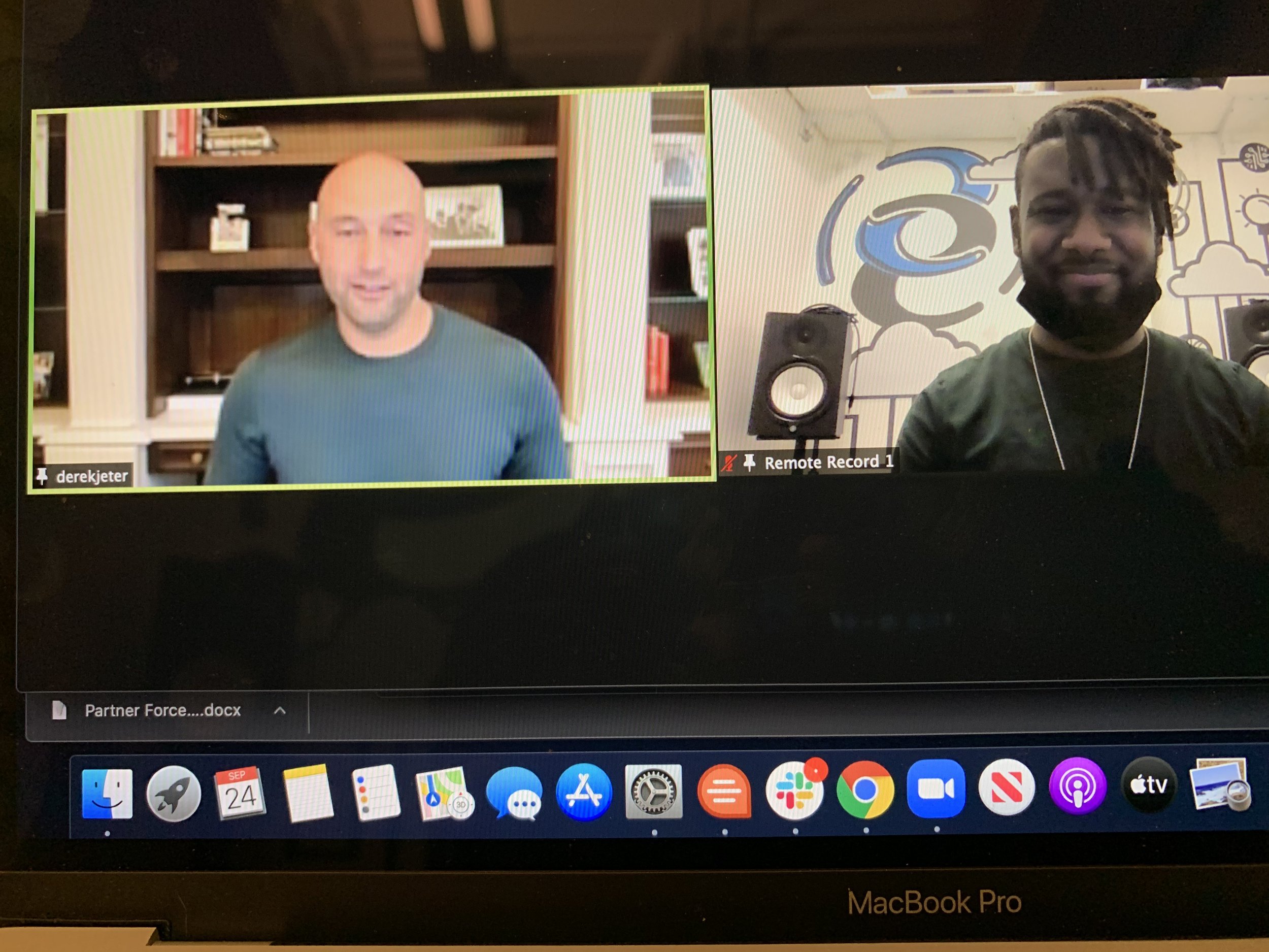 A Zoom video conference showing two men in separate locations. The man on the left has a bald head and is smiling, sitting in front of a wooden bookshelf with books and framed pictures. The man on the right has dark skin and dreadlocks, sitting in a 