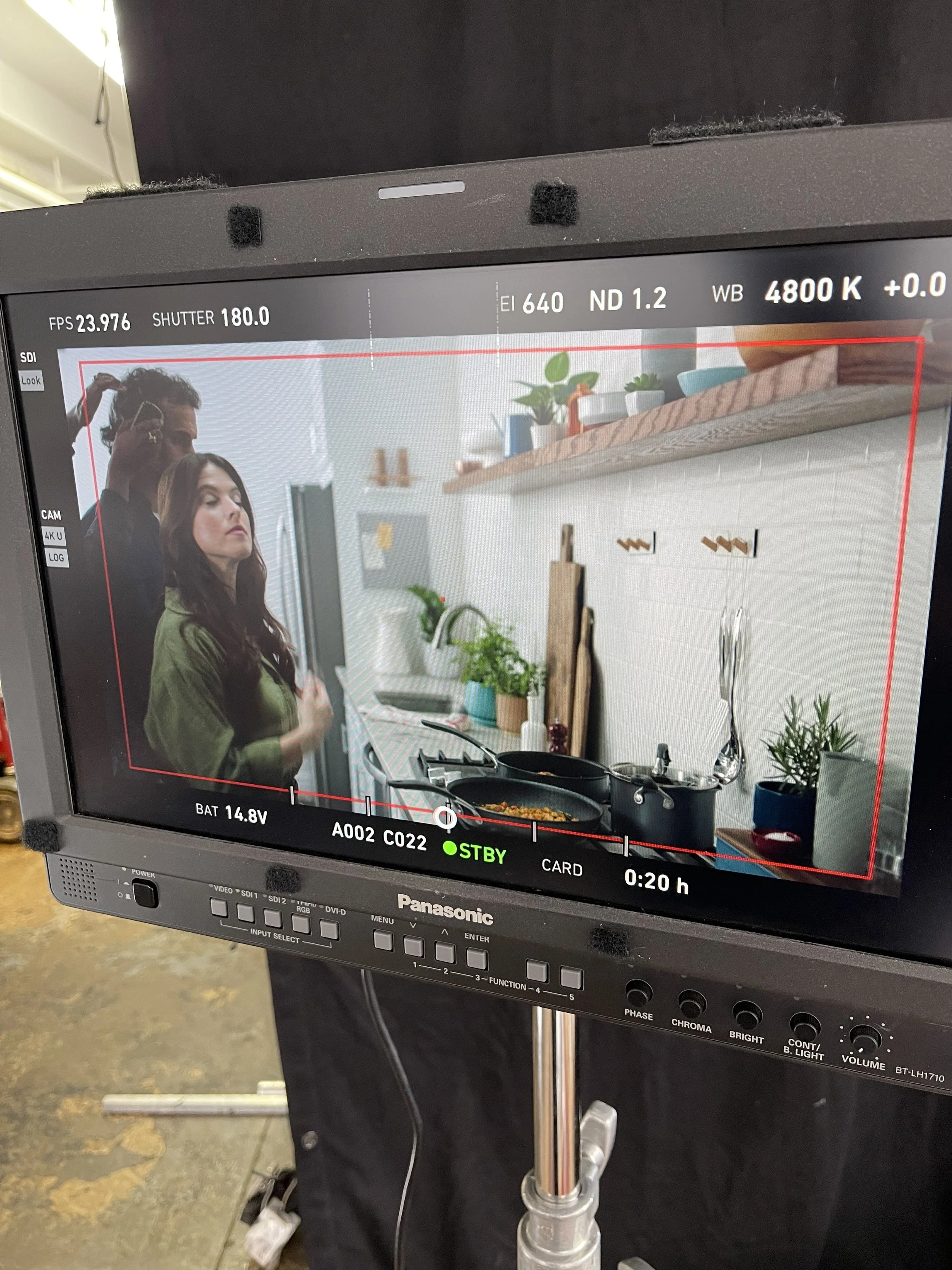 A woman in a green jacket stands in a modern kitchen, with a man in the background styling her hair on a film or video shoot, as seen through the monitor screen.
