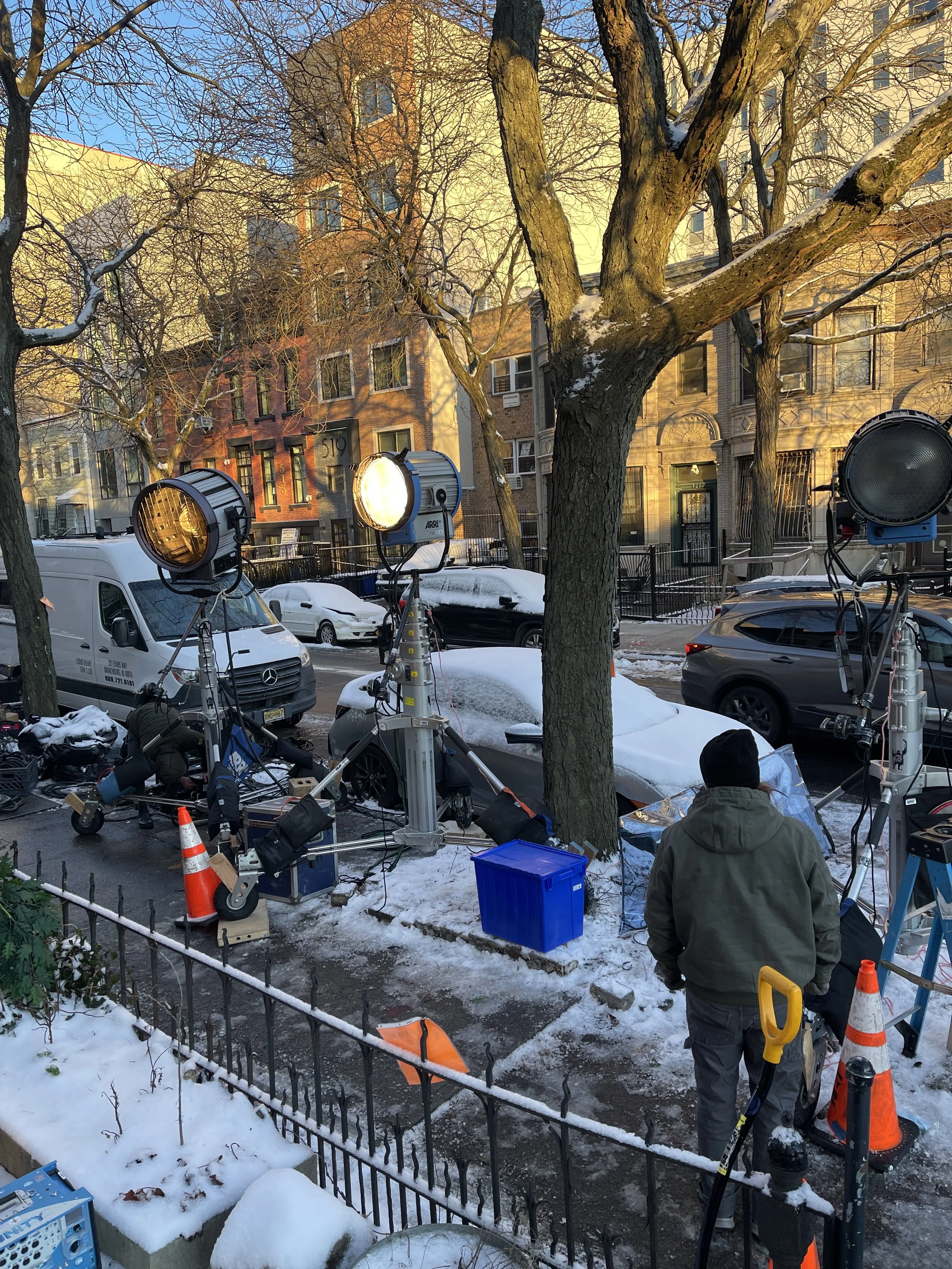 Filming crew setting up lights on a snowy city street with parked cars and apartment buildings in the background.
