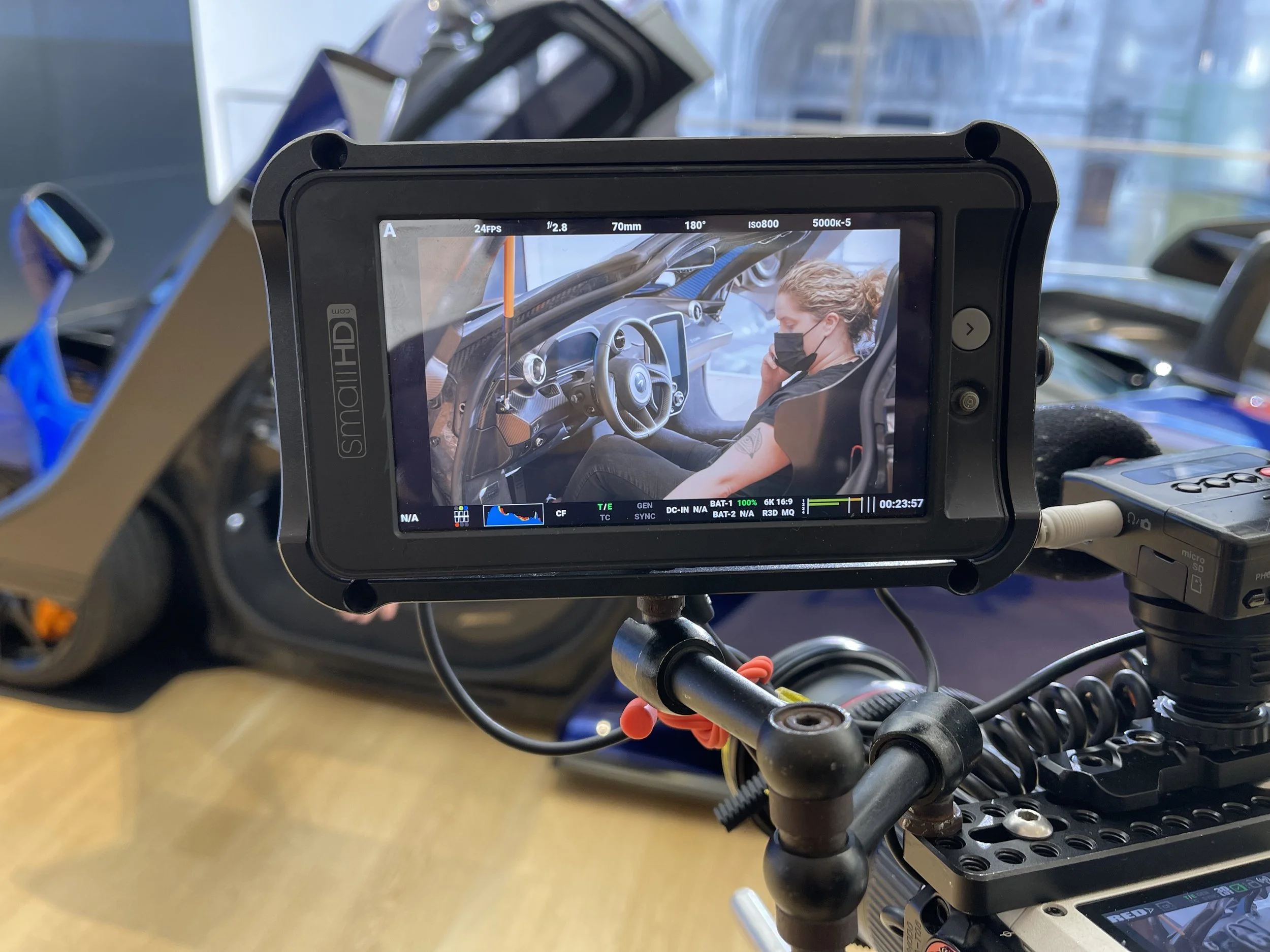 Close-up of a camera monitor on a film set showing a woman wearing a black face mask sitting inside a race car, with the car's interior visible in the background.