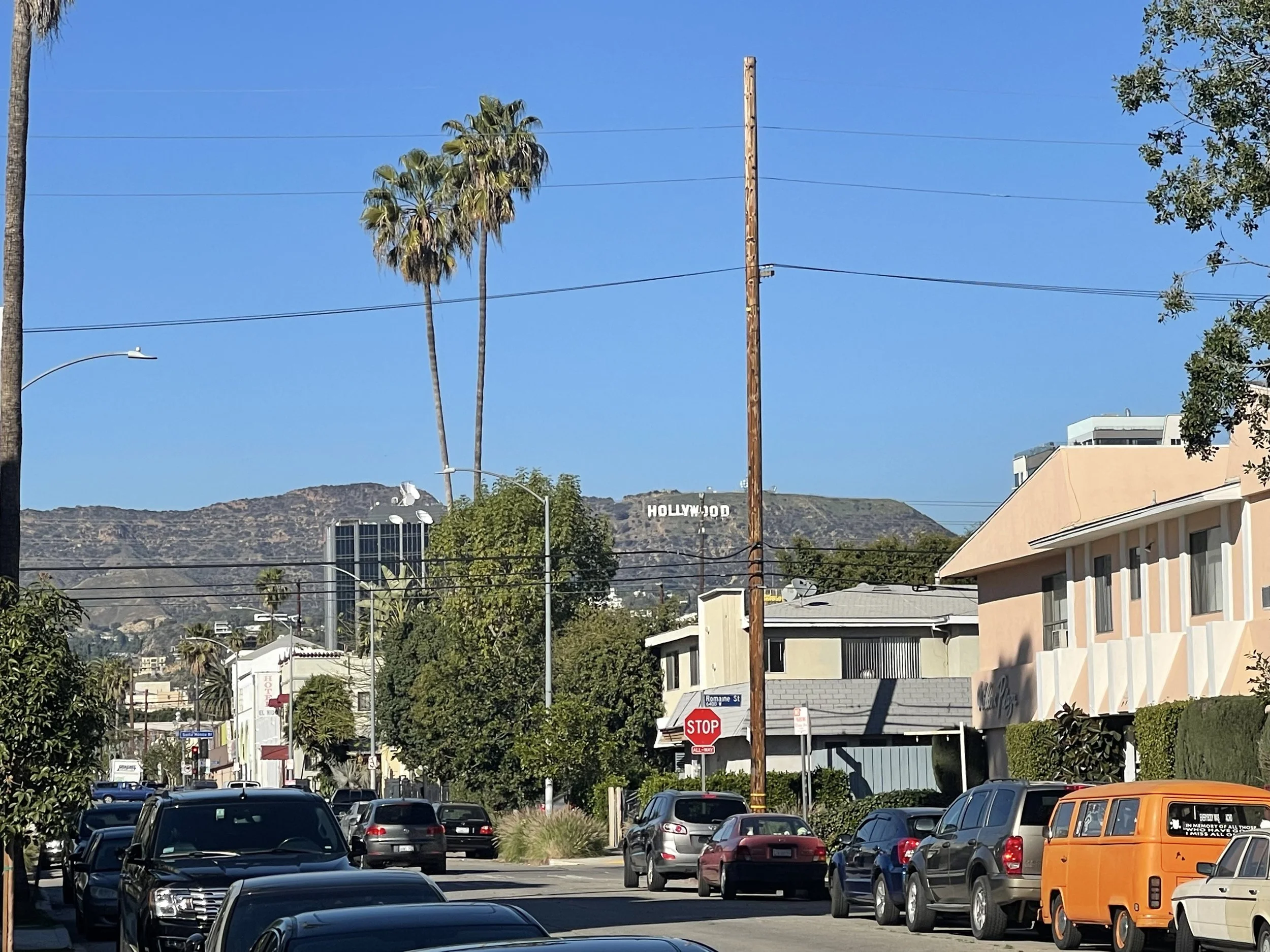 Street scene in Los Angeles with cars parked on the side, palm trees, residential buildings, and the Hollywood sign on the hillside in the background.