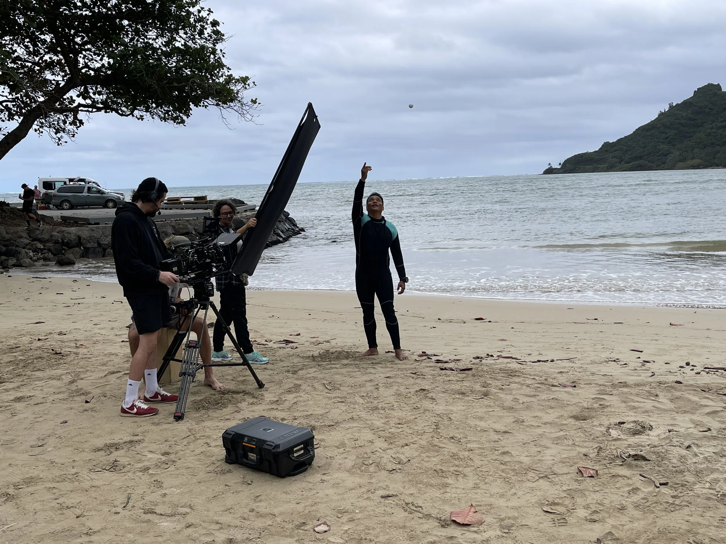 A person in a wetsuit throws a ball on a beach while a camera crew films the scene, overlooking the ocean with cloudy skies and a hill in the background.