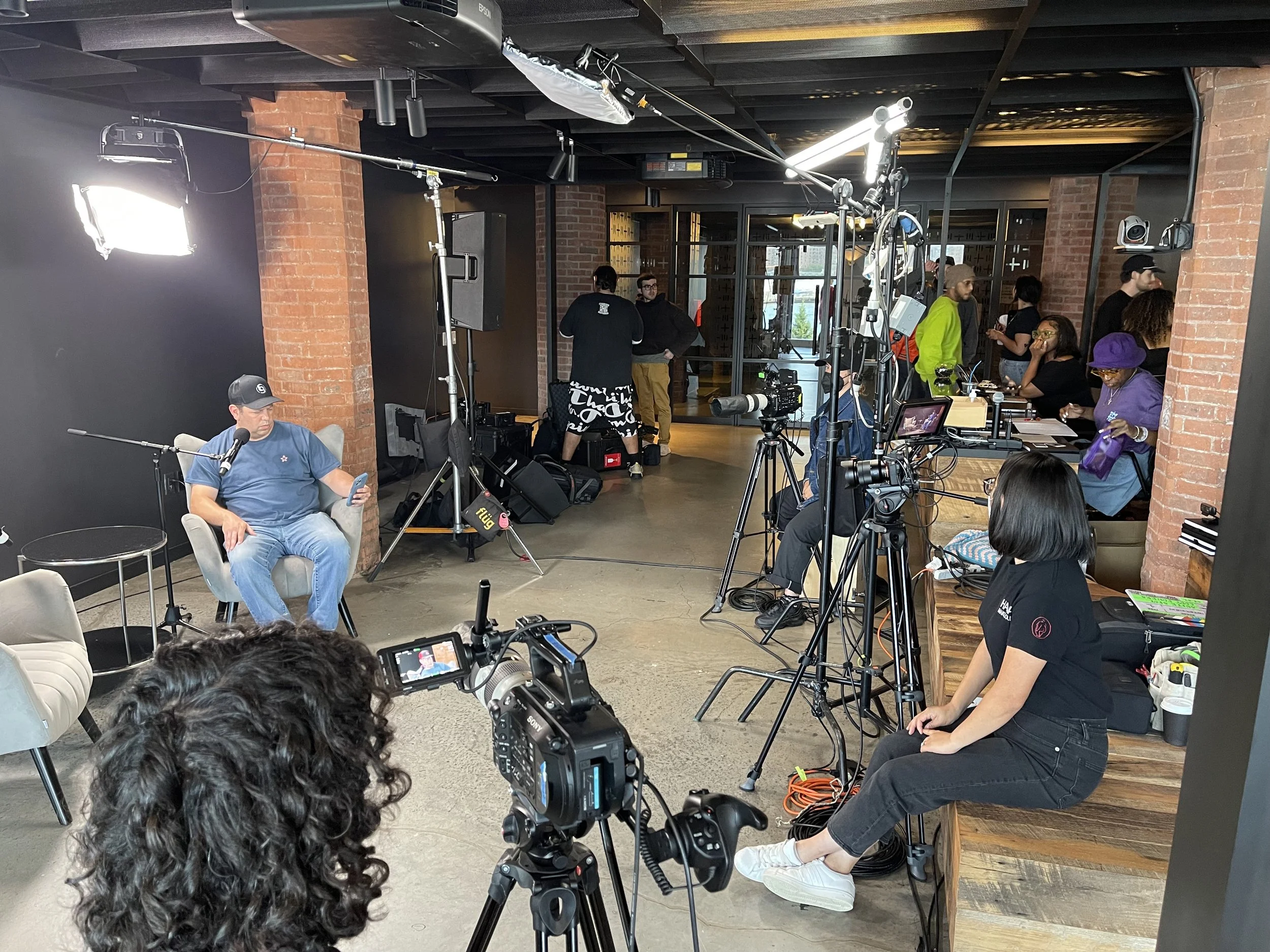 A behind-the-scenes view of a film or video shoot in a modern brick-walled space with multiple cameras, lighting equipment, crew members, and a man sitting in a chair with a microphone, preparing for a recording or interview.