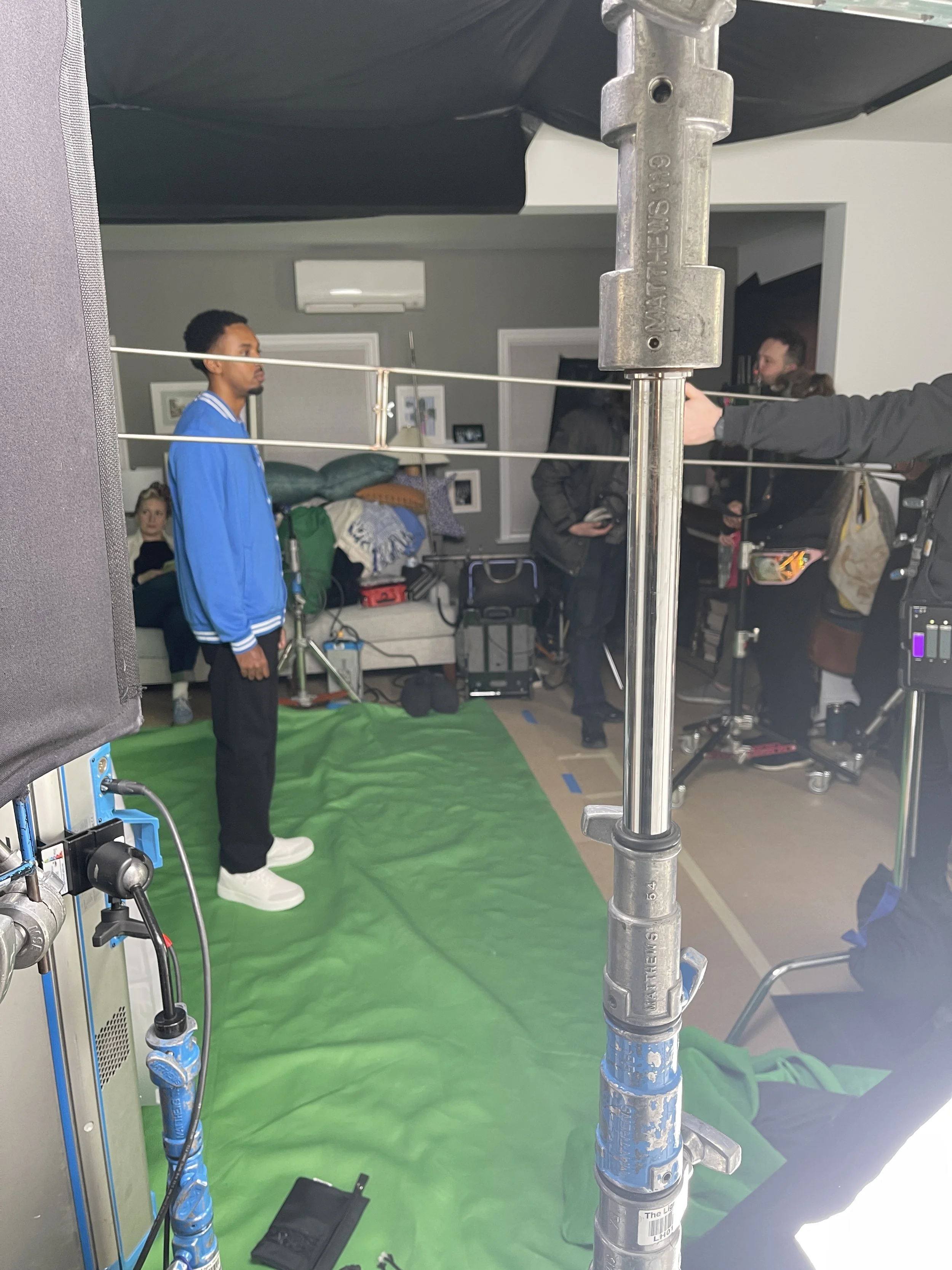 A scene from a film or photo shoot setup showing a man in a blue jacket standing on a green screen floor, with crew members and equipment around him in a living room.