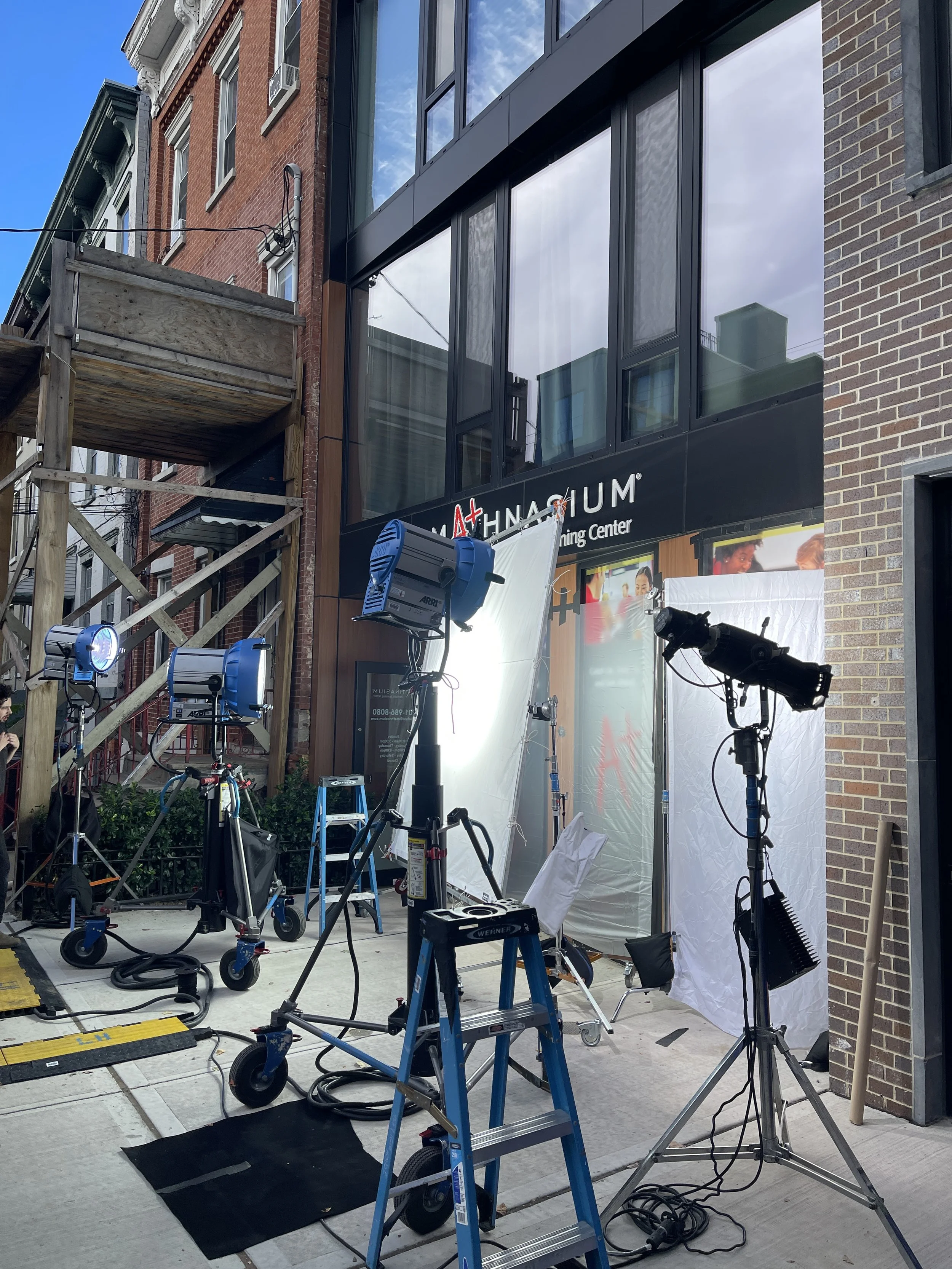 Outdoor film or photo shoot setup with lighting equipment, white screens, and a backdrop in front of a building with signs for 'Mathnasium.'