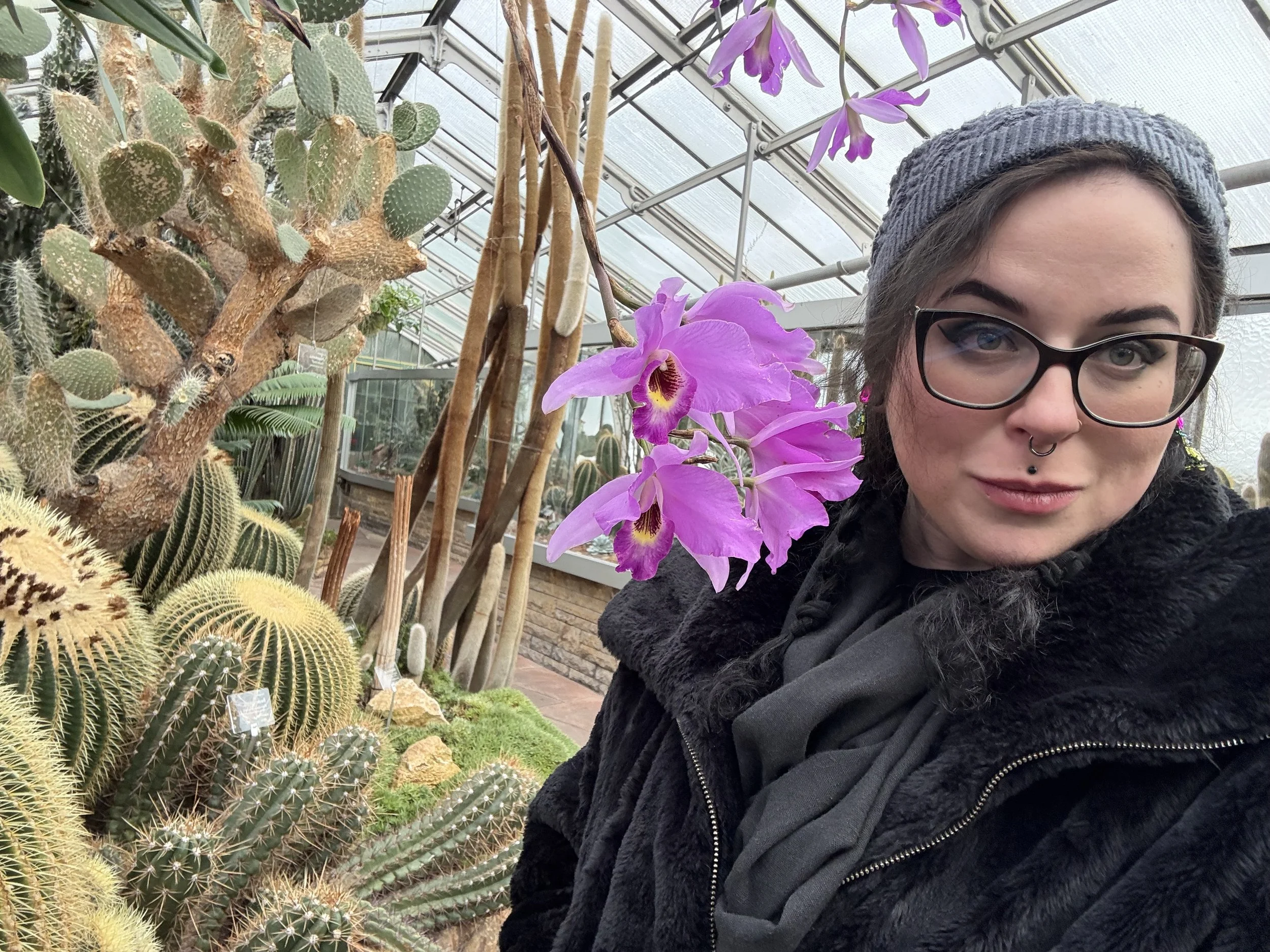 A woman with glasses and a gray beanie taking a selfie in a greenhouse, surrounded by various cacti and pink orchids.