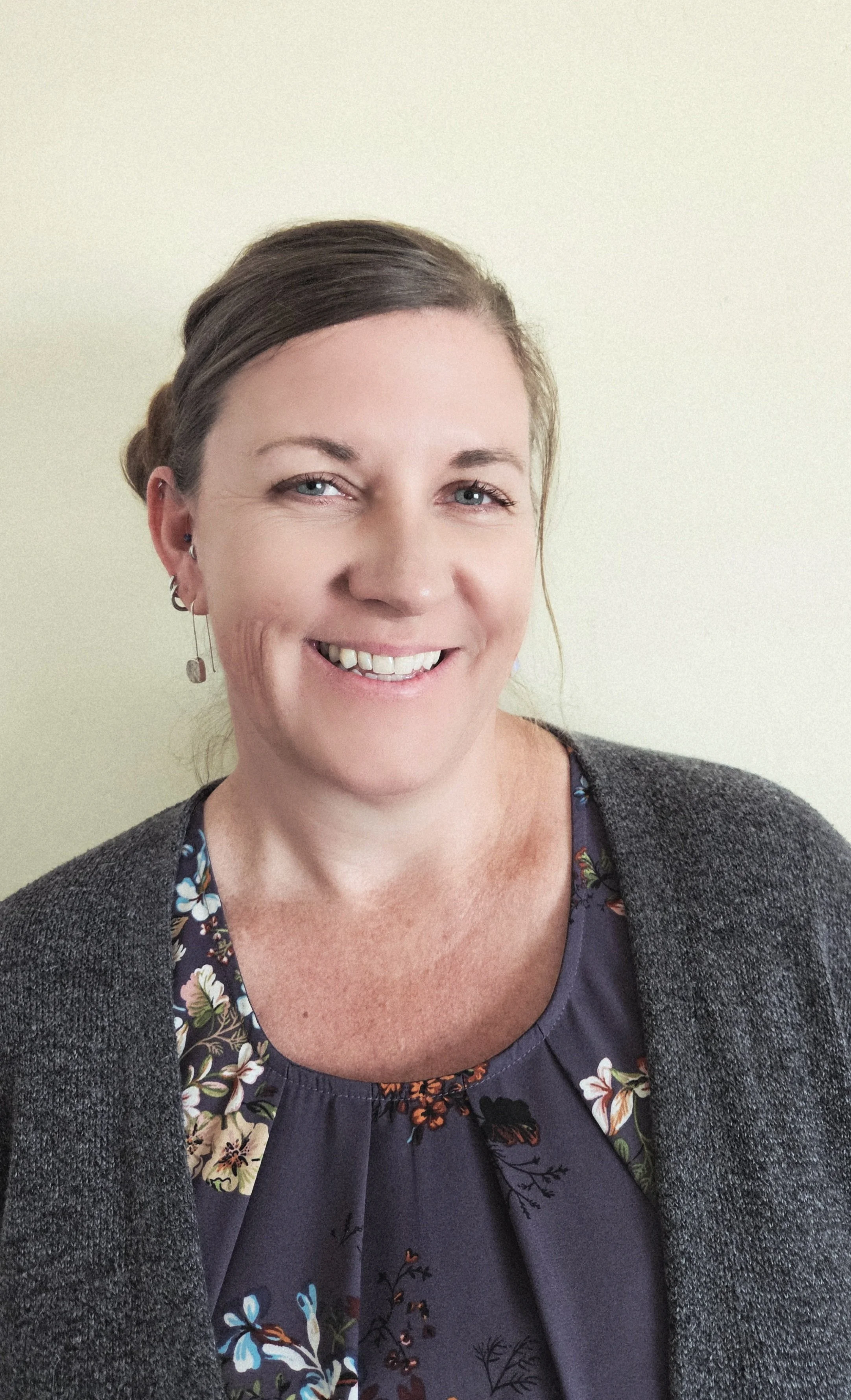 A woman smiling, wearing a gray cardigan over a floral blouse, with light-colored background.