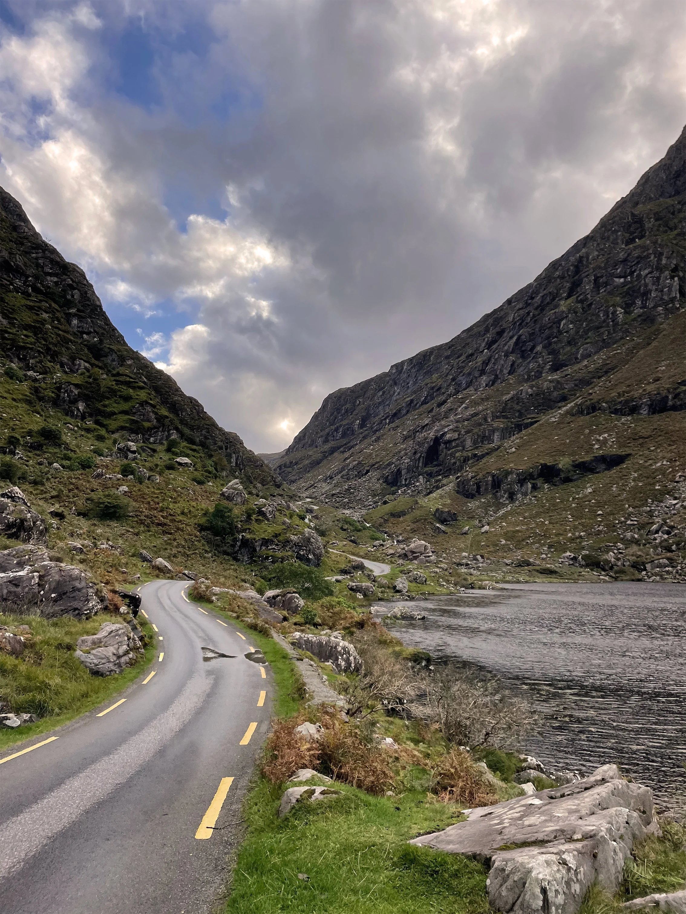 A winding road beside a lake in a mountainous landscape with rocky hills and a cloudy sky.