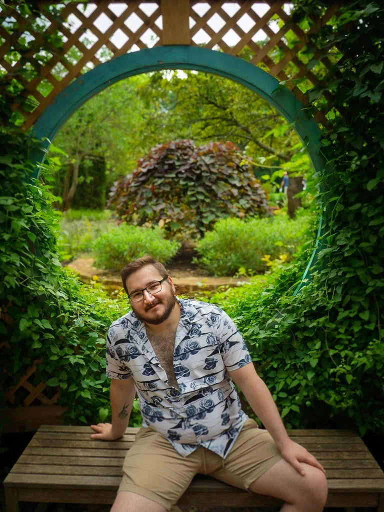 A man with glasses and a beard sitting on a wooden bench in a garden. He is wearing a patterned button-up shirt and beige shorts, surrounded by lush green foliage and a lattice archway.