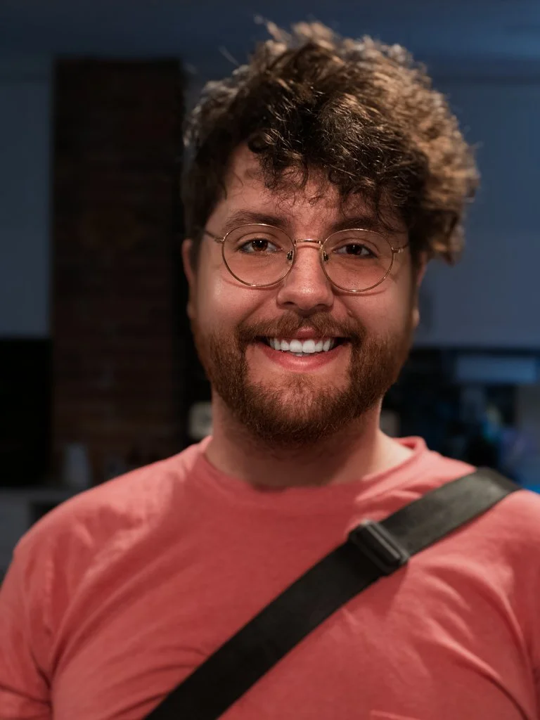 Smiling man with glasses and a beard, wearing a pink shirt and a black strap, indoors with dim lighting.