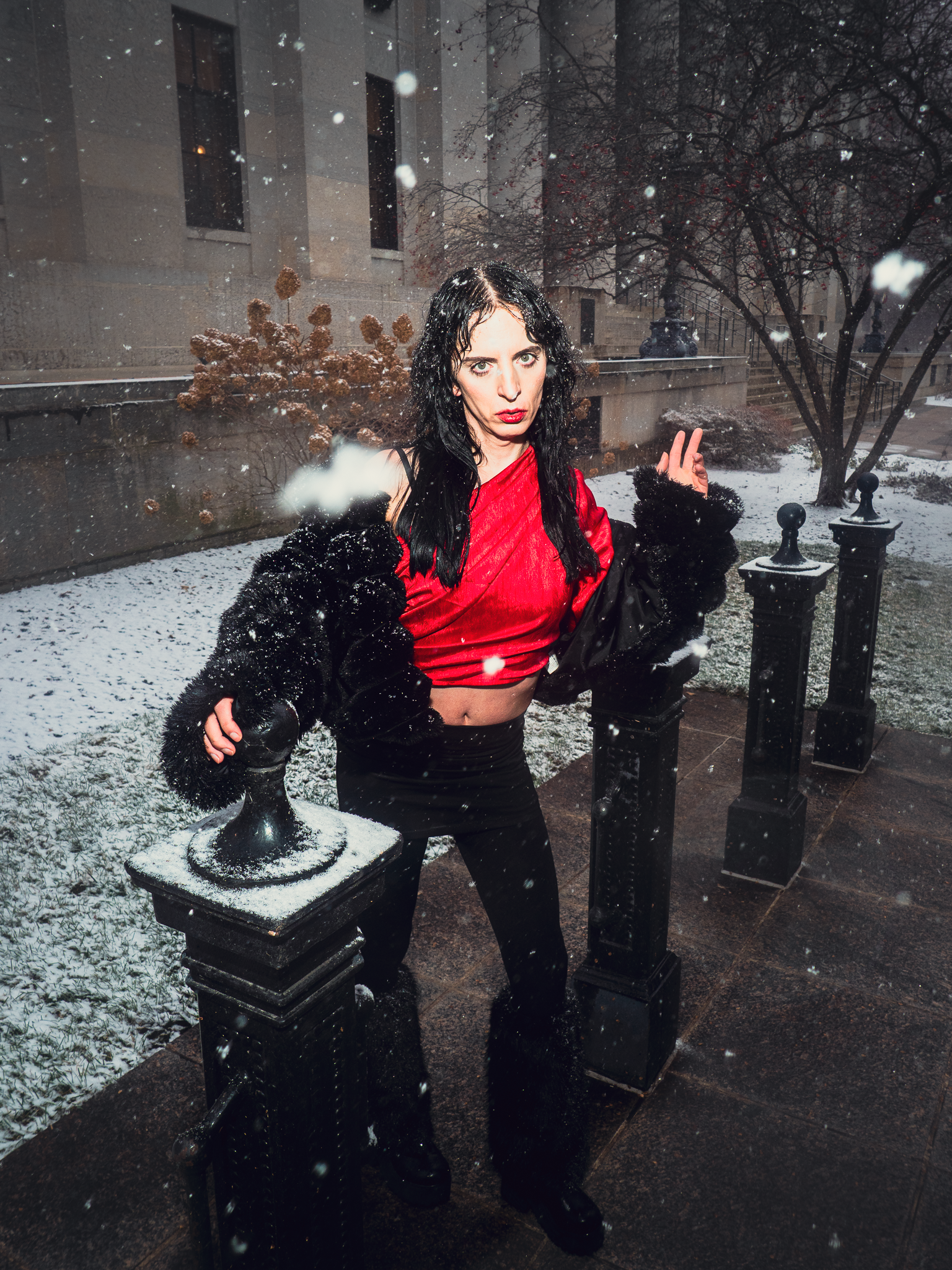 A woman with long black hair, wearing a red crop top and black furry jacket, stands outdoors in a snowy environment with her hands against black fence posts, looking directly at the camera.